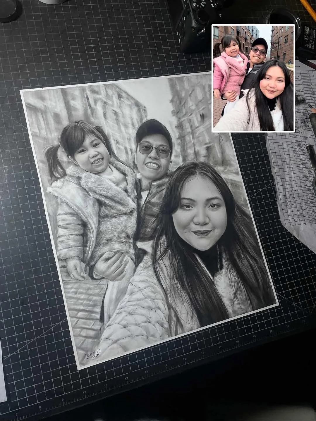 Black and white pencil drawing of a family of three, with a woman, man, and young girl, on an artist's work table with a photograph of the same family in the corner.