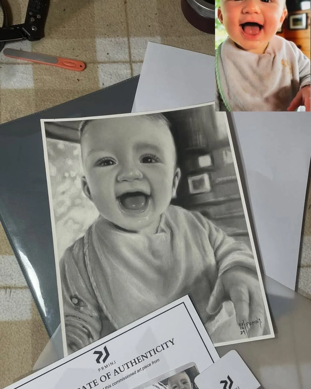 A black-and-white realistic portrait of a smiling baby with a wide-open mouth, wearing a shirt with suspenders, and pointing at the camera.