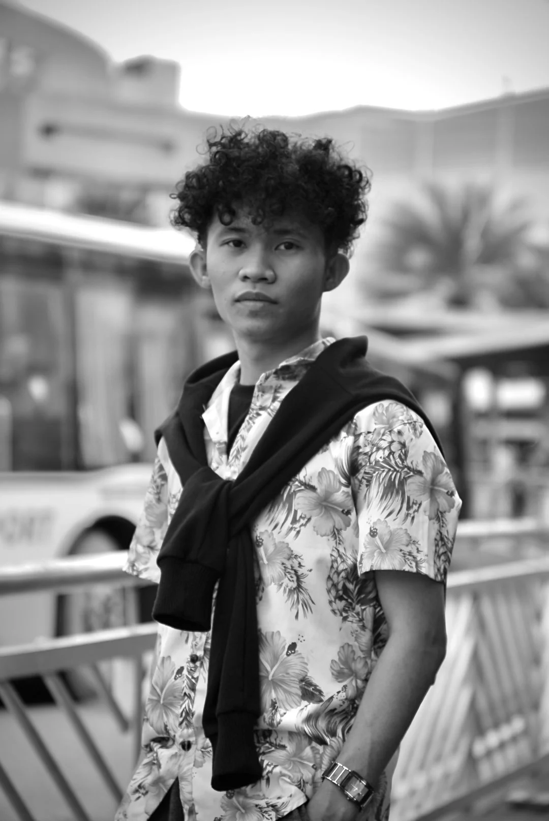 A young man with curly hair wearing a tropical print shirt and a watch, standing outdoors with a railing and cityscape in the background.