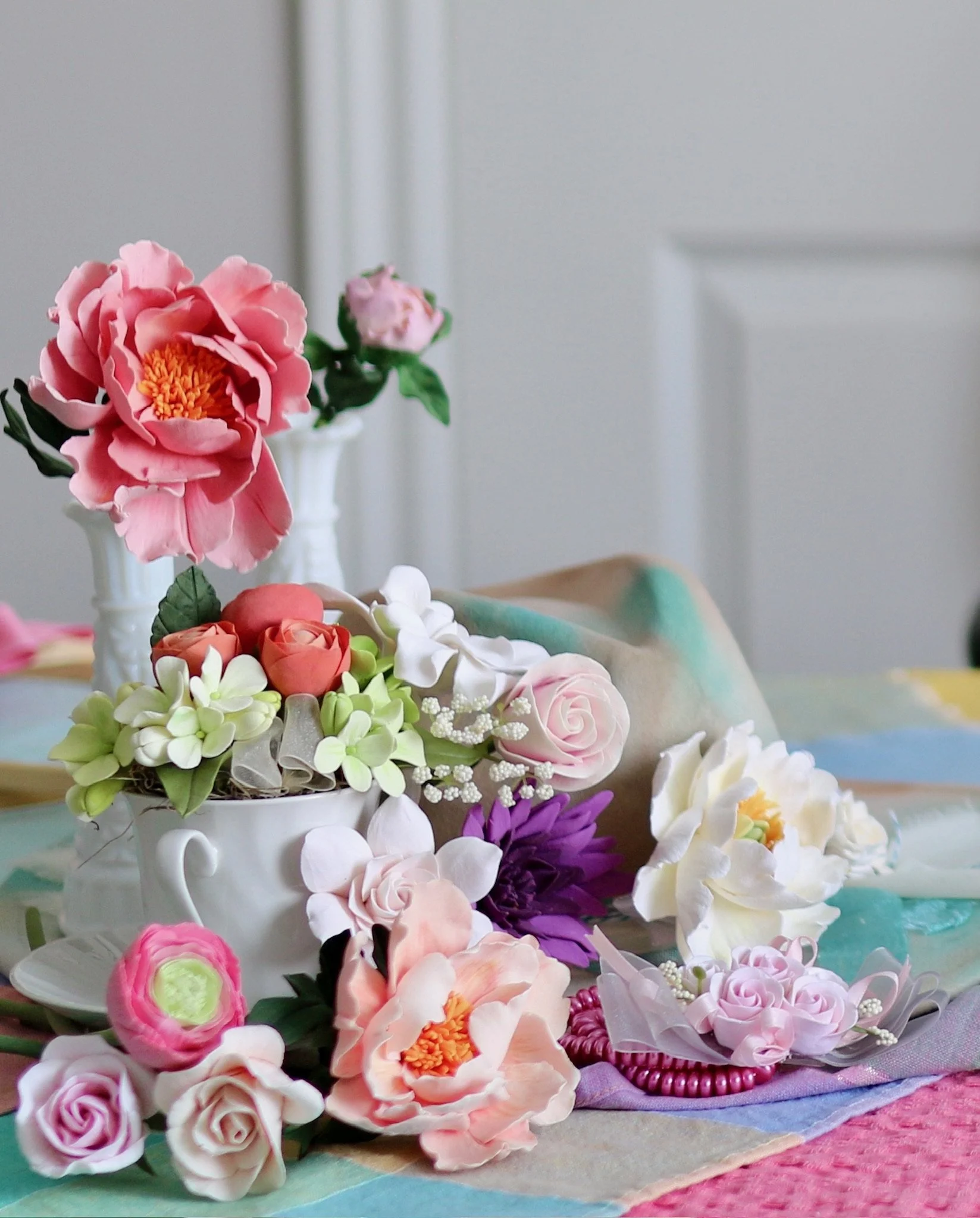A table display of stunning clay flowers arranged in vases and a cup, including pink, white, purple, and orange flowers. Each bloom is created by hand, petal by petal. Custom colours, flower varieties and orders welcomed.