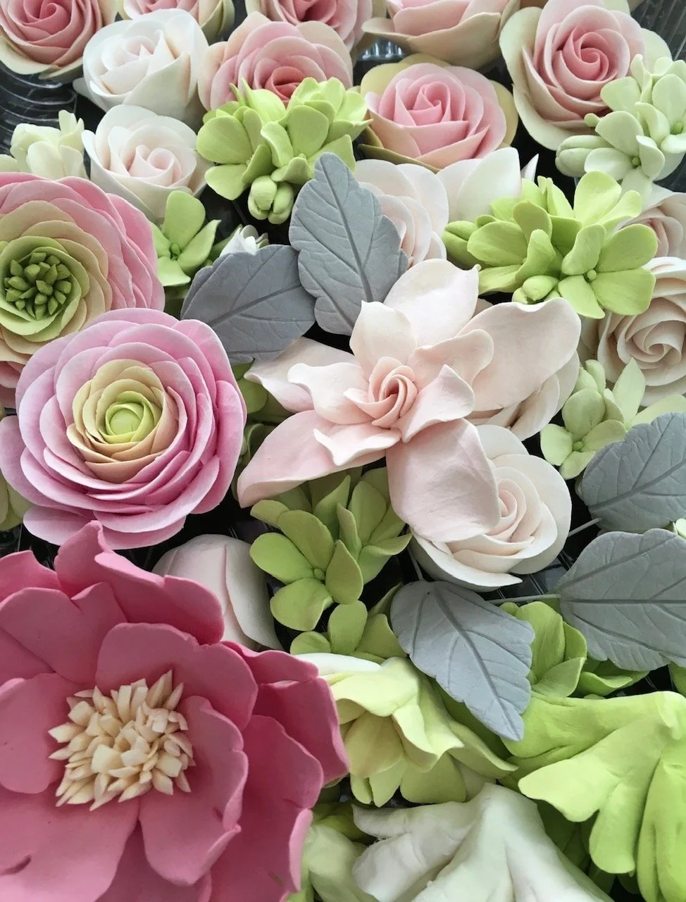 An assortment of pastel-coloured, everlasting clay flowers including roses, peonies, and hydrangea blossoms with green and gray dusty miller leaves.