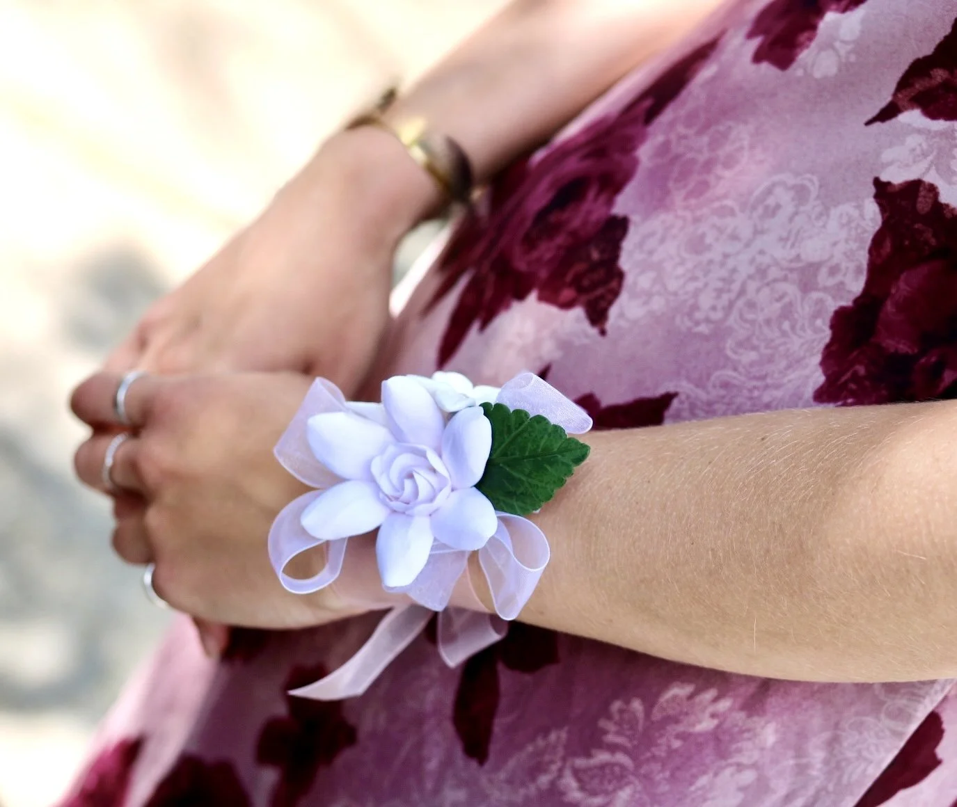 Close-up of a young woman's arm wearing a white floral wrist corsage made of a clay gardenia and green leaves with a pink ribbon, over a pink and burgundy floral dress.