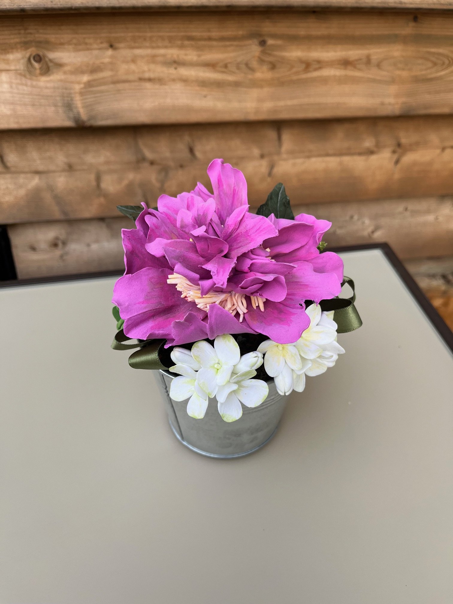 An open and bloomed magenta peony in a small flower arrangement accented with white flowers with green leaves, sitting on a glass table with a wooden shiplap background.