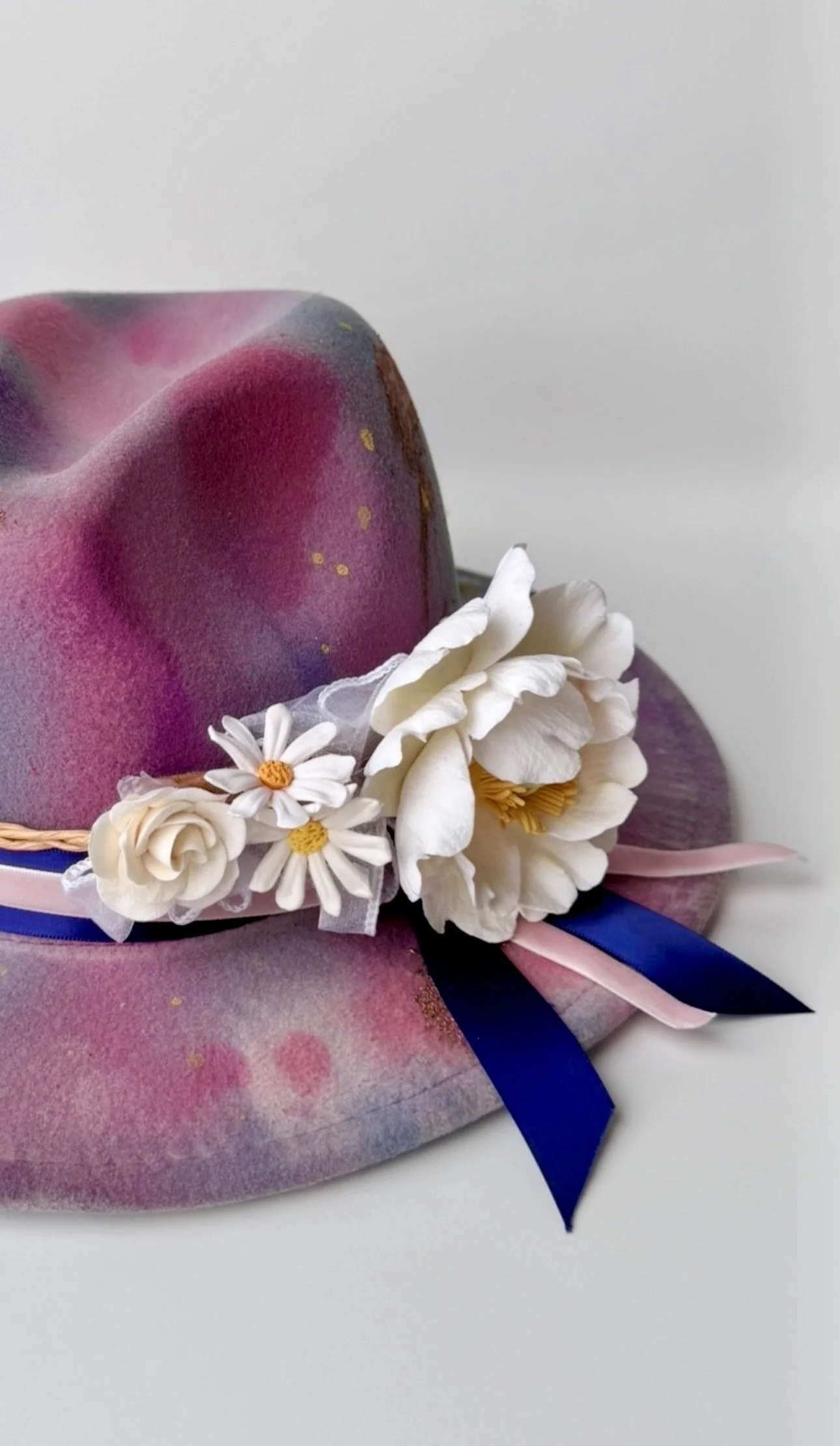 Clay flowers that include white peonies, daisies and roses wrapped around a one-of-a-kind, watercolour painted wide-brim felt hat, decorated with blue and pink ribbons, against a white background.