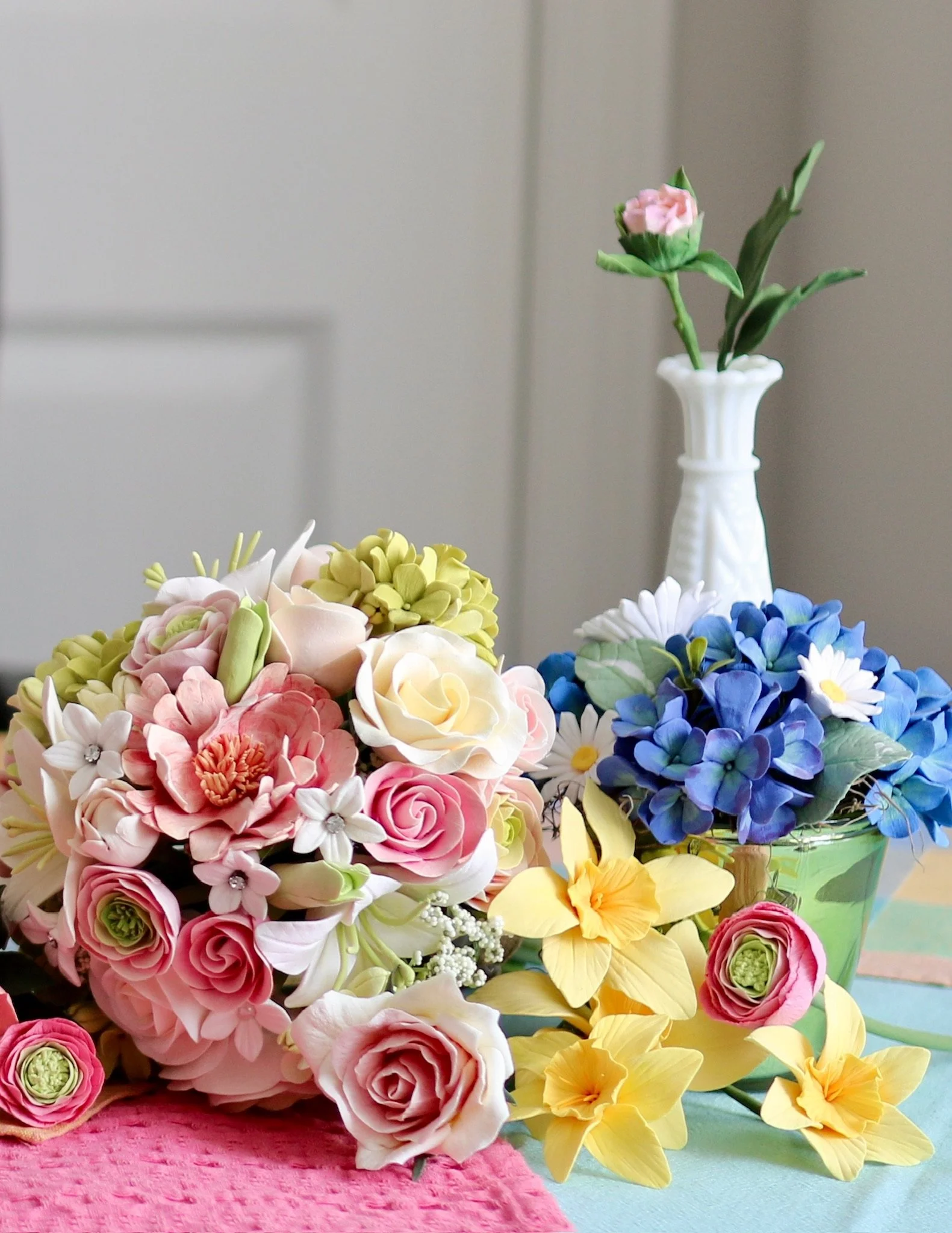Arrangement of colourful, everlasting clay flowers including pink roses, white roses, blue hydrangeas, yellow daffodils, and other one-of-a-kind looms on a table with a pink cloth and a lightcoloured background.