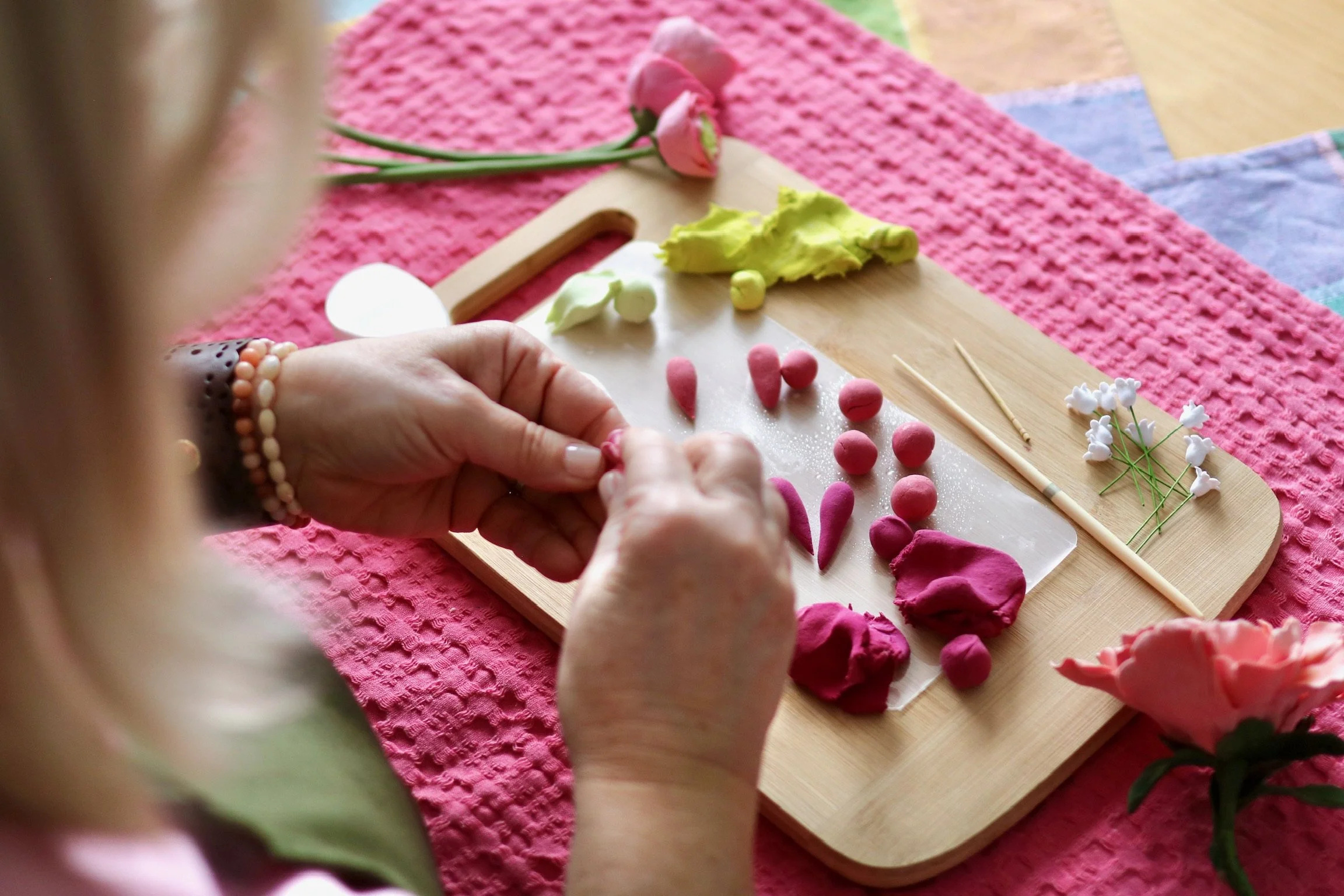 Over the shoulder view of clay flower artist, Tracey Paul using air-dry clay, making balls to mold into colourful flower petals. Working on a solid board, surrounded by flowers and art tools.