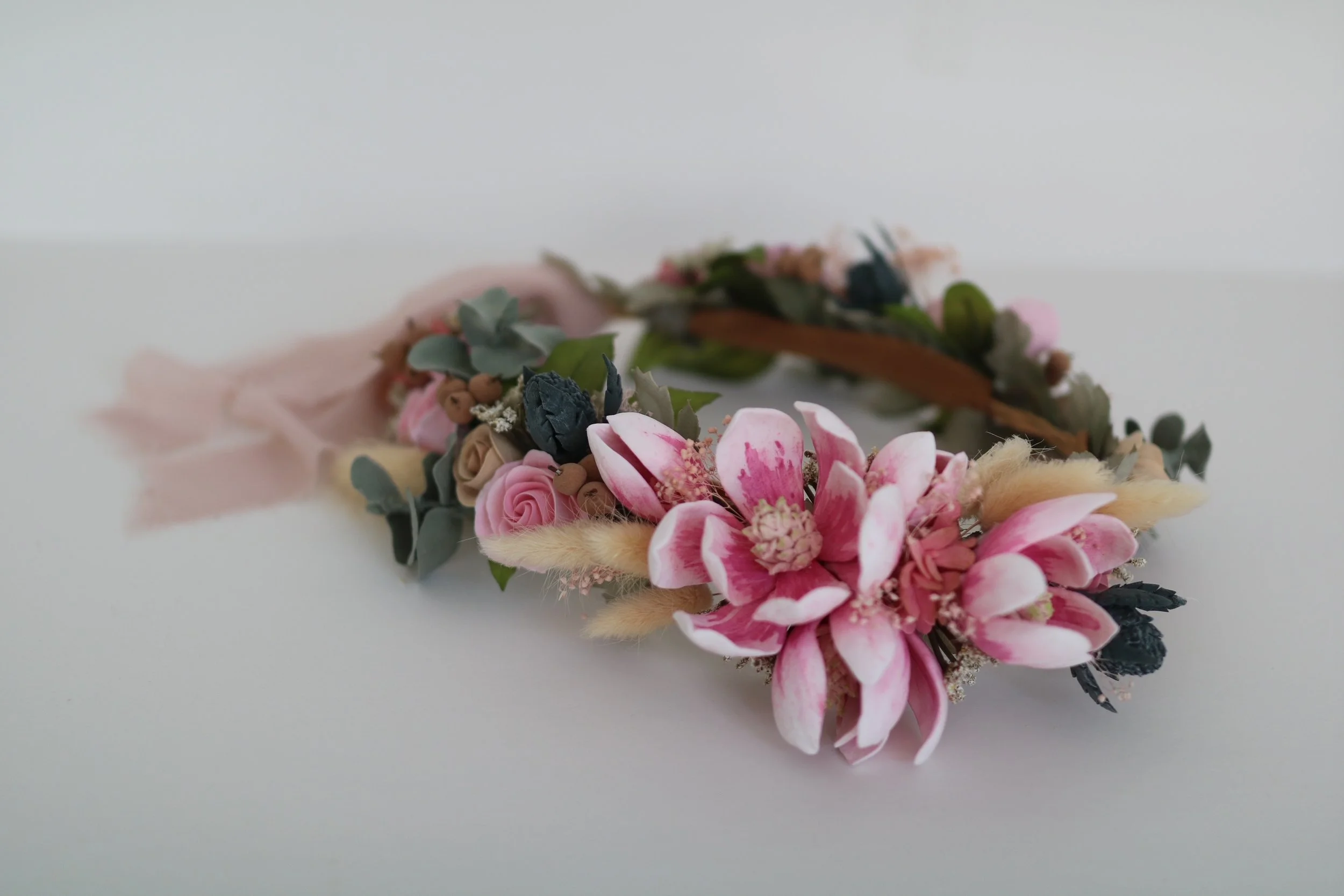 A floral headband crown with pink and white magnolia and roses, teal thistles and beige accent roses along with green leaves, resting on a white surface.