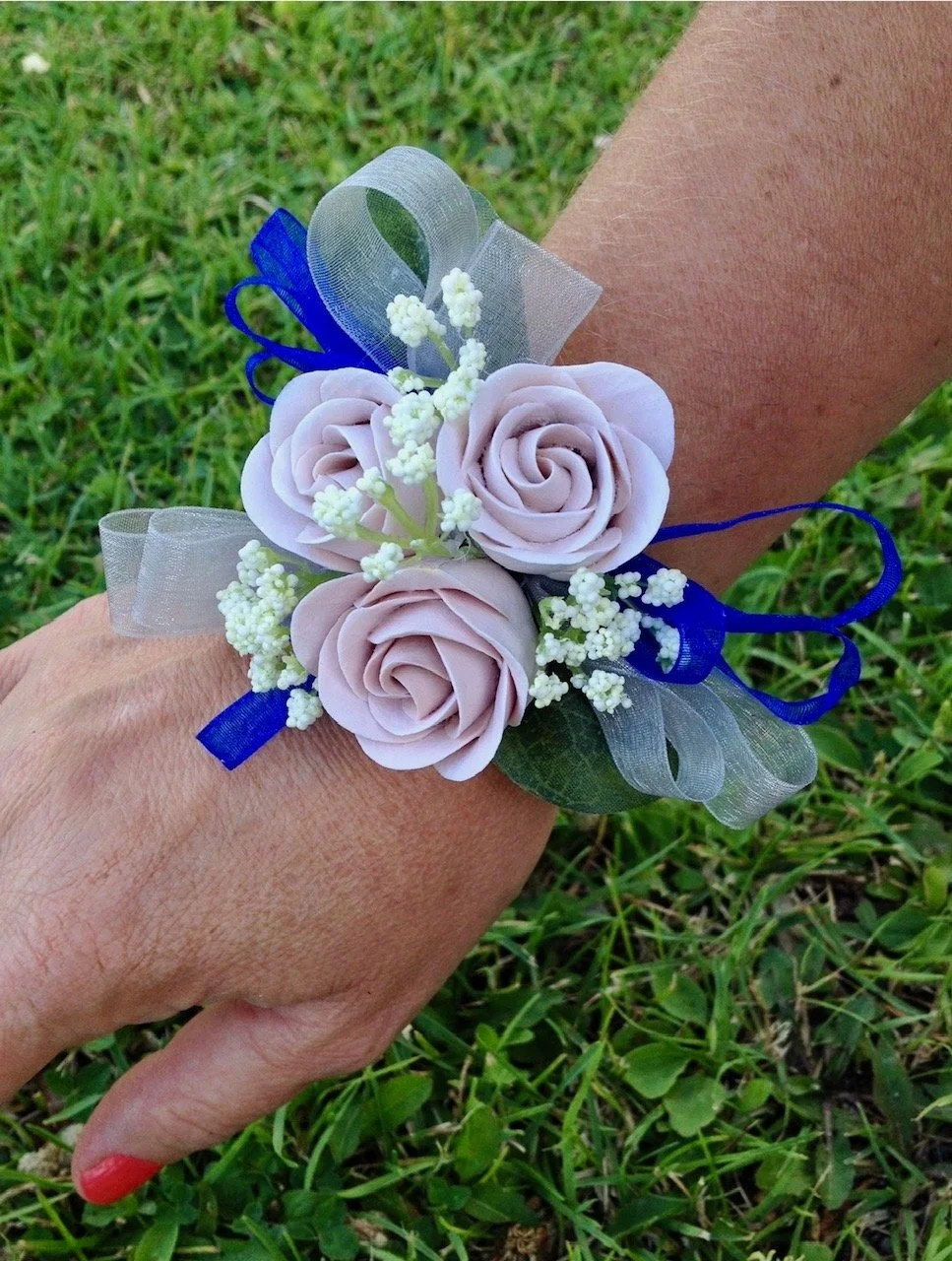 A floral wrist corsage with three pale pink roses, white baby's breath, green leaves, with blue and silver ribbons, worn by a person with coral nail polish, against a backdrop of green grass.