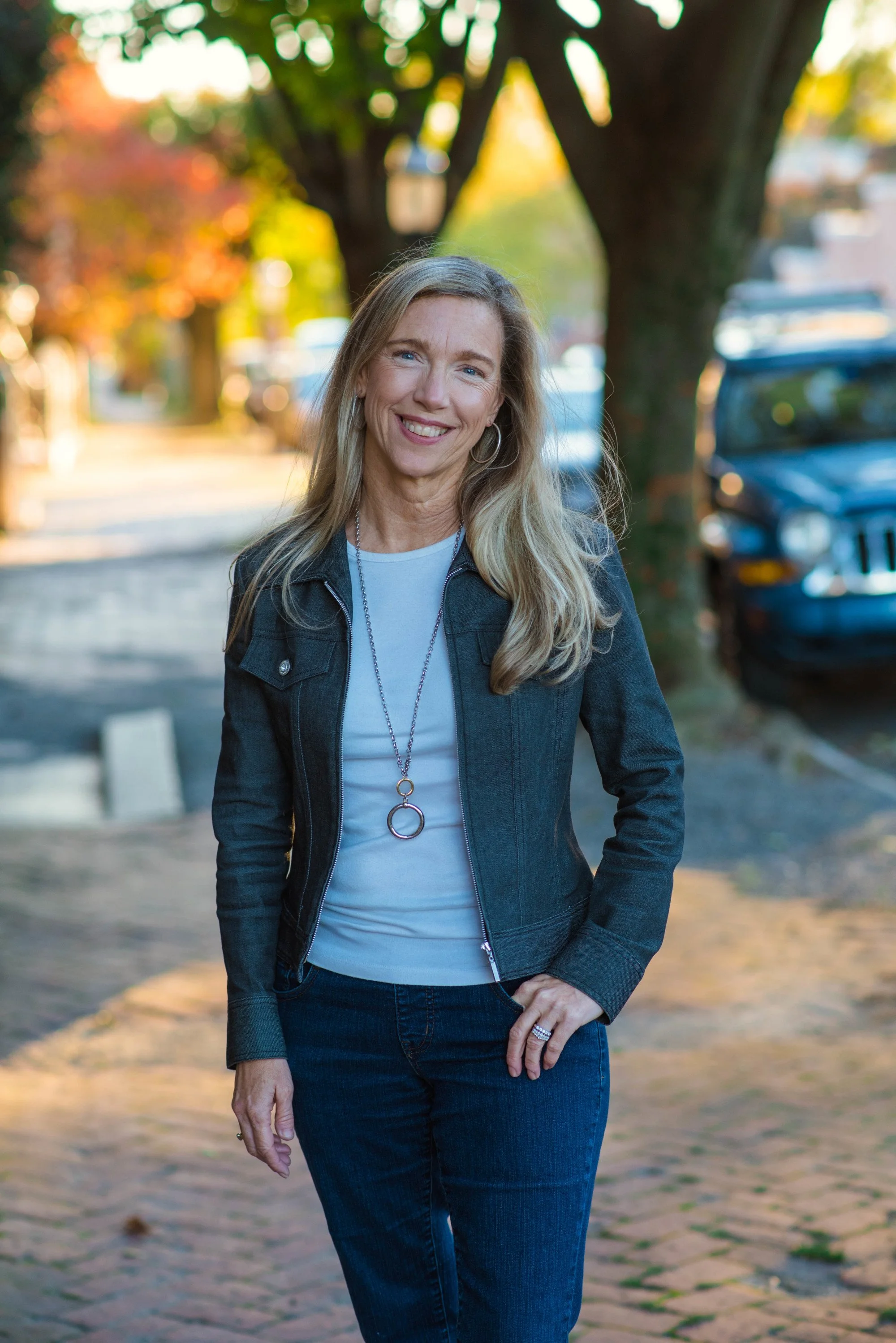 A woman with long blonde hair smiling and standing outdoors on a brick path with trees and cars in the background during autumn.