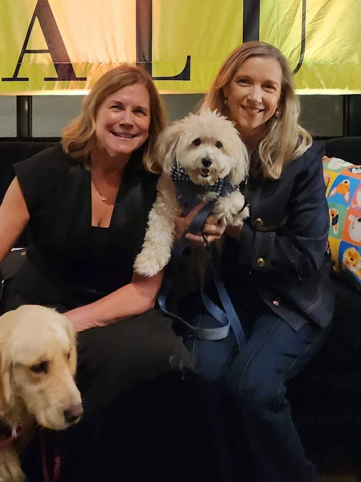 Two women smiling and holding a small white fluffy dog with a bandana. A yellow lab is partially visible in the foreground, and a colorful pillow in the background.