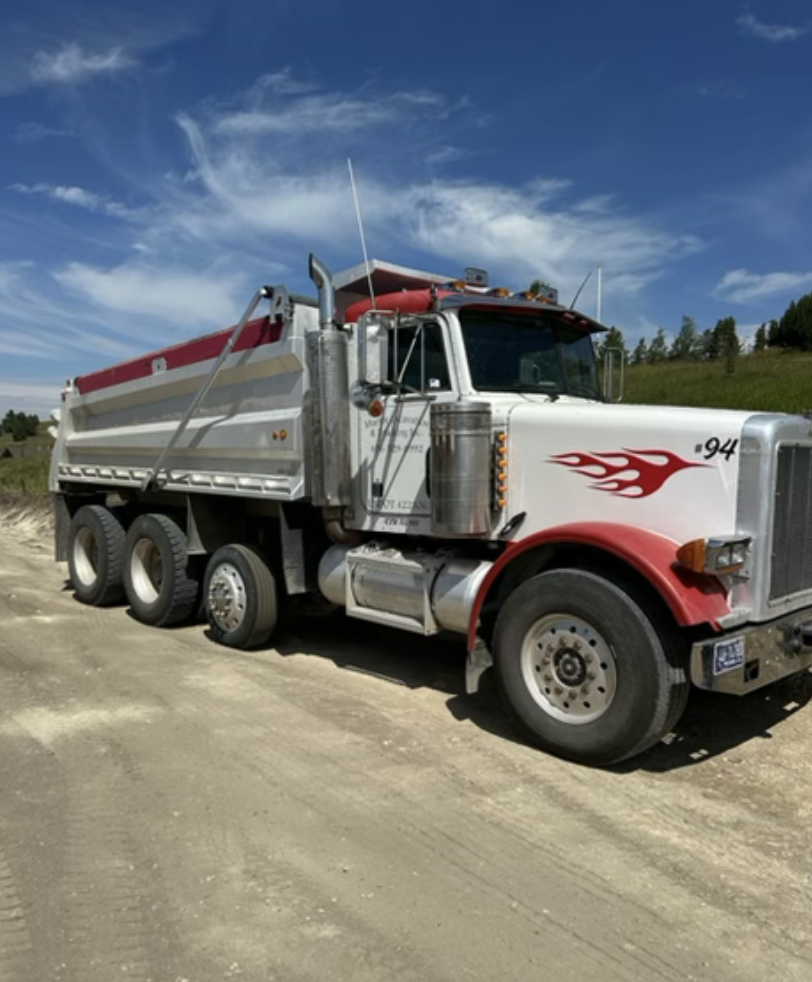 A white dump truck with a flame decal on the front and red accents parked on a dirt road under a partly cloudy sky.
