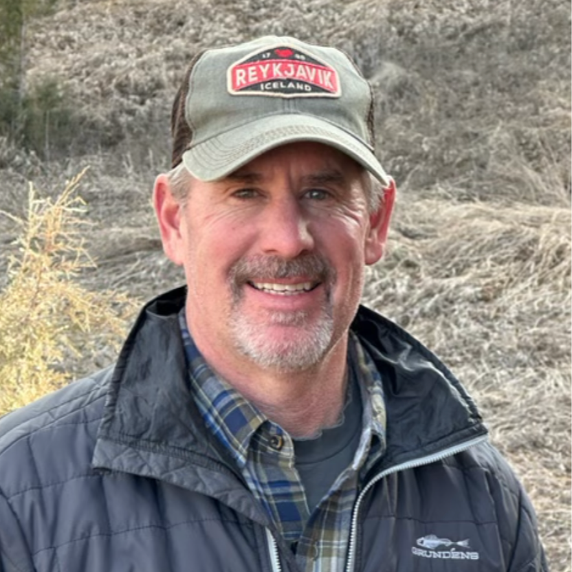 A man smiling outdoors wearing a baseball cap with "Reykjavik Iceland" embroidered, a dark jacket, and a plaid shirt, with a dry, grassy landscape in the background.