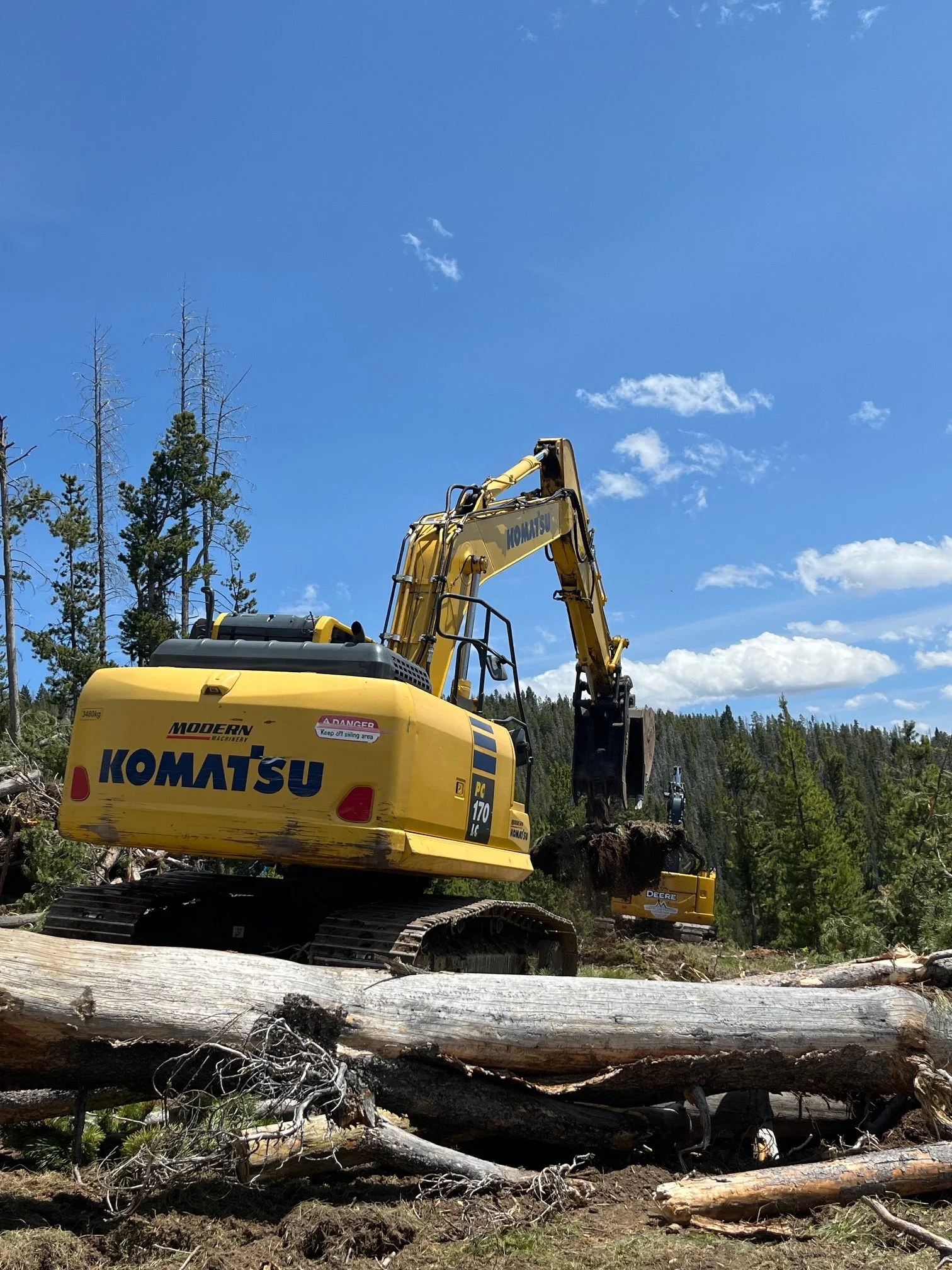 A yellow Komatsu excavator is digging and moving logs in a forested area with scattered trees under a clear blue sky.