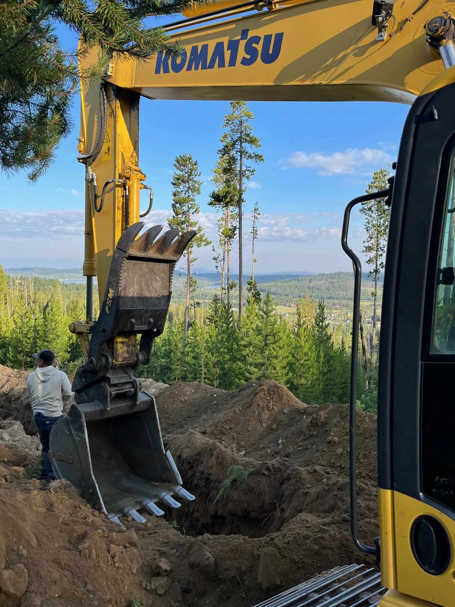 Construction site with a yellow Komatsu excavator and a worker near a deep trench in a wooded area with pine trees and a scenic view of the landscape and mountains in the background.