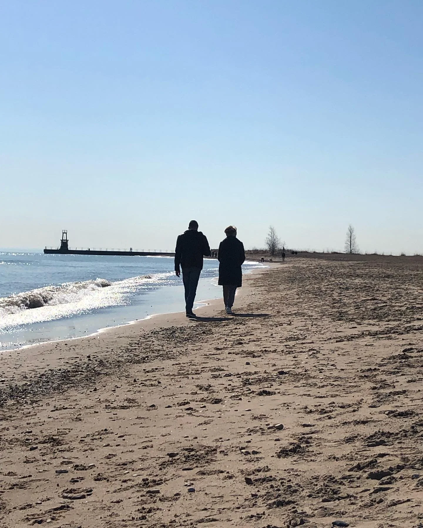 Two people walk along the sandy beach near the water, with a pier in the distance under a clear blue sky.