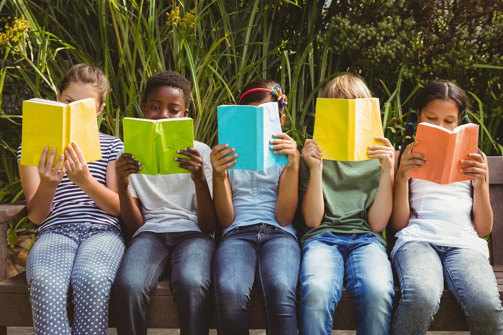 Five children sitting on a bench reading books, surrounded by green plants, outdoors.