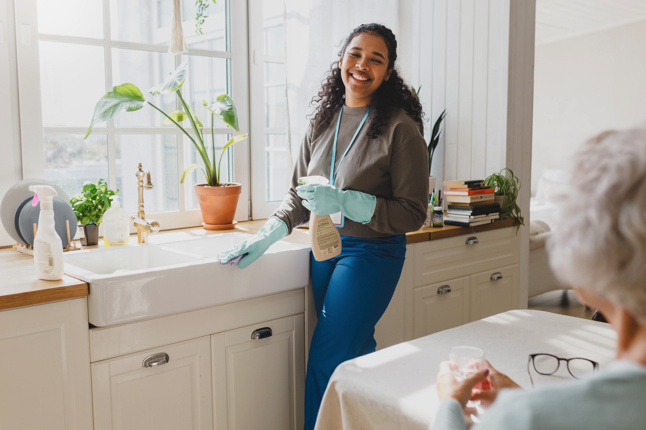 A woman wearing gloves cleaning a kitchen sink with a spray bottle, while a person with glasses observes at the table.