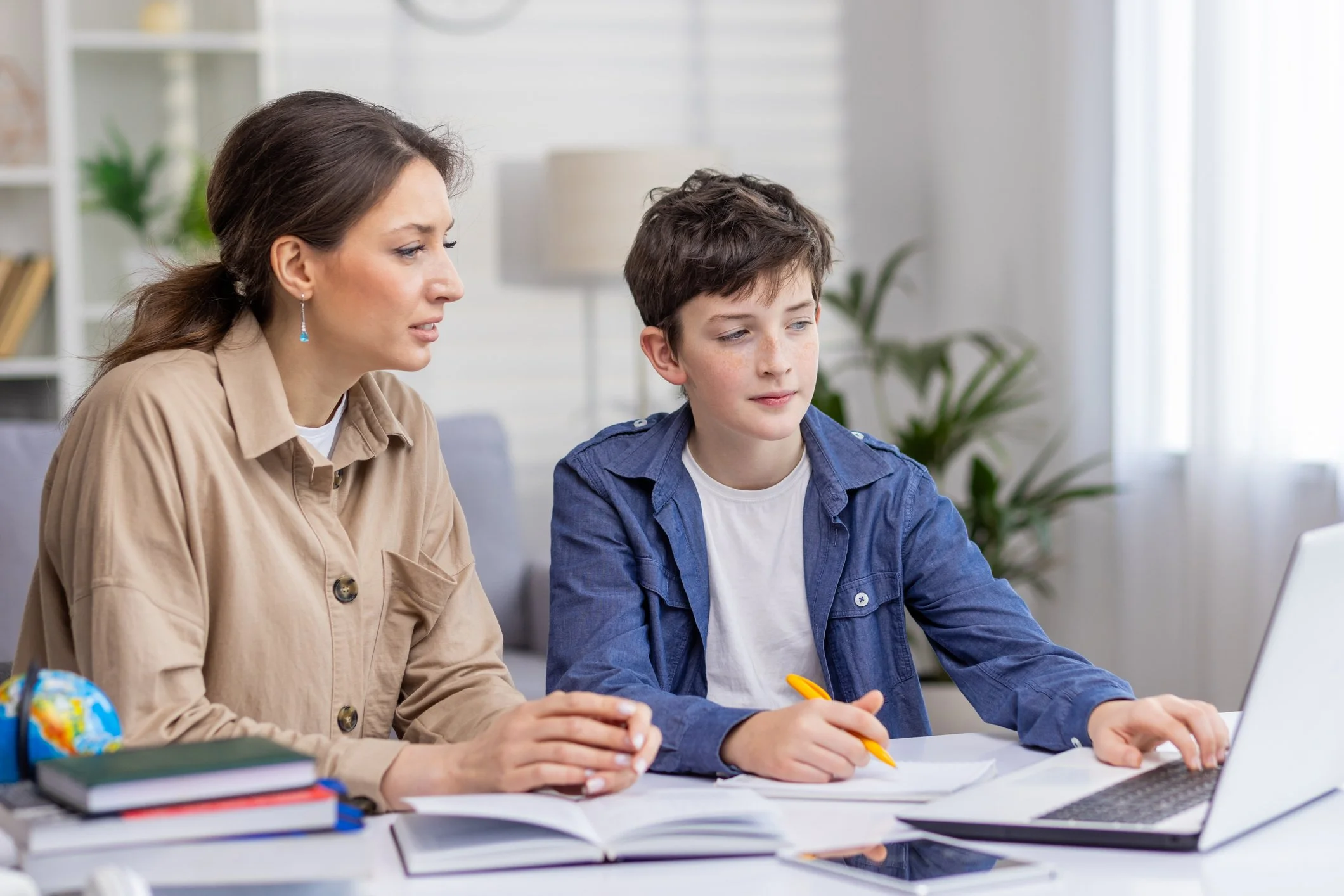 A woman and a boy sitting at a desk with books, a globe, and a laptop, working together on a school assignment.