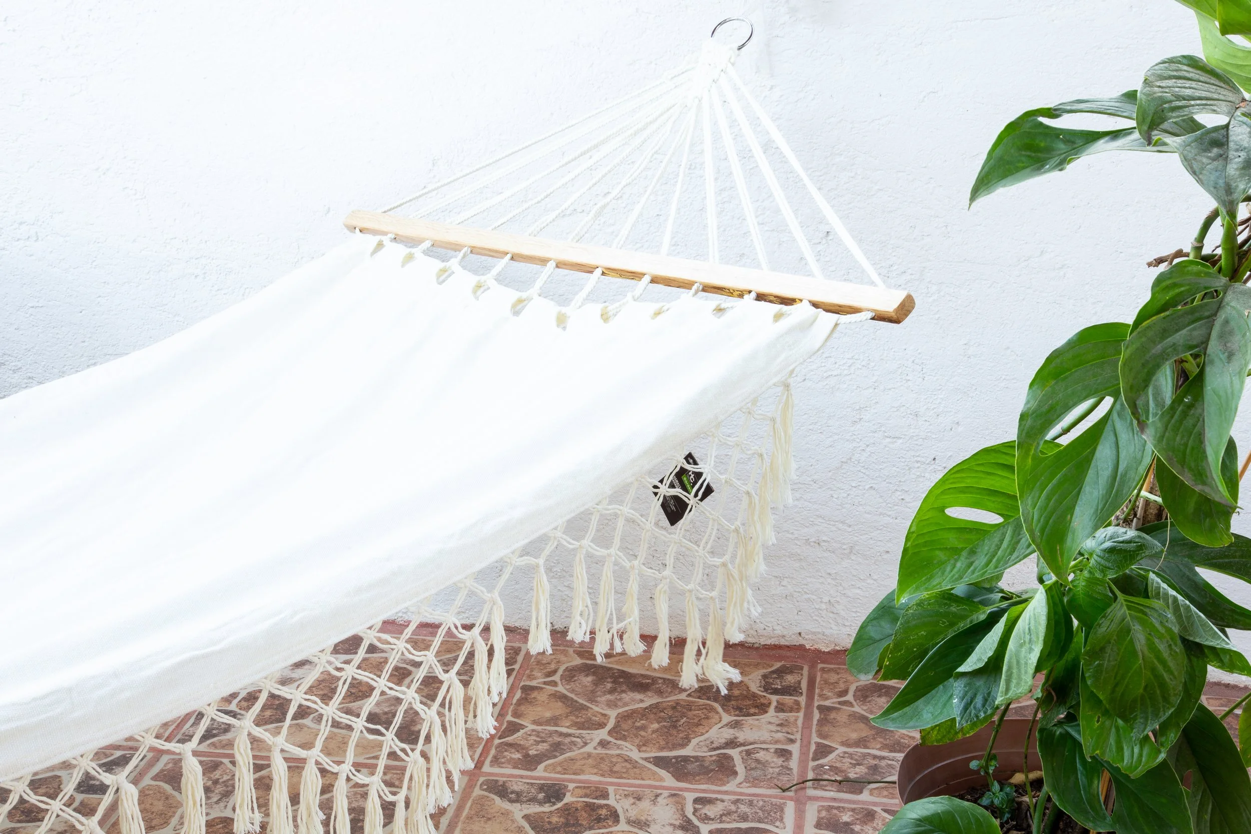 Hamaca blanca colgada en una pared blanca, rodeada de plantas verdes, en un piso de baldosas de cerámica.