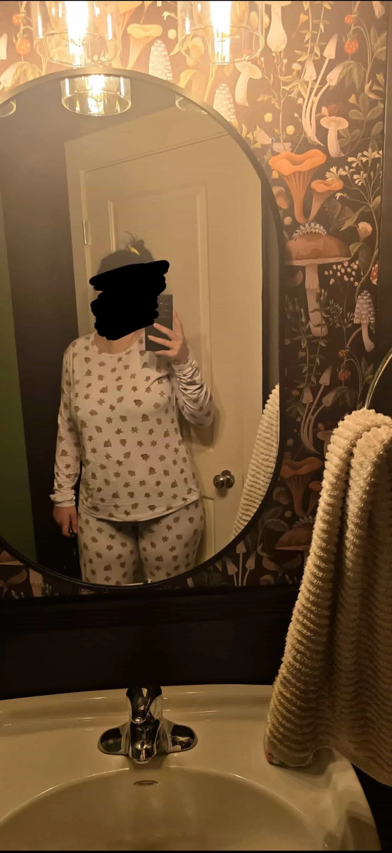 A person in pajamas with a star and holiday pattern taking a selfie in a bathroom mirror, with towels hanging on a rack and floral wallpaper in the background.