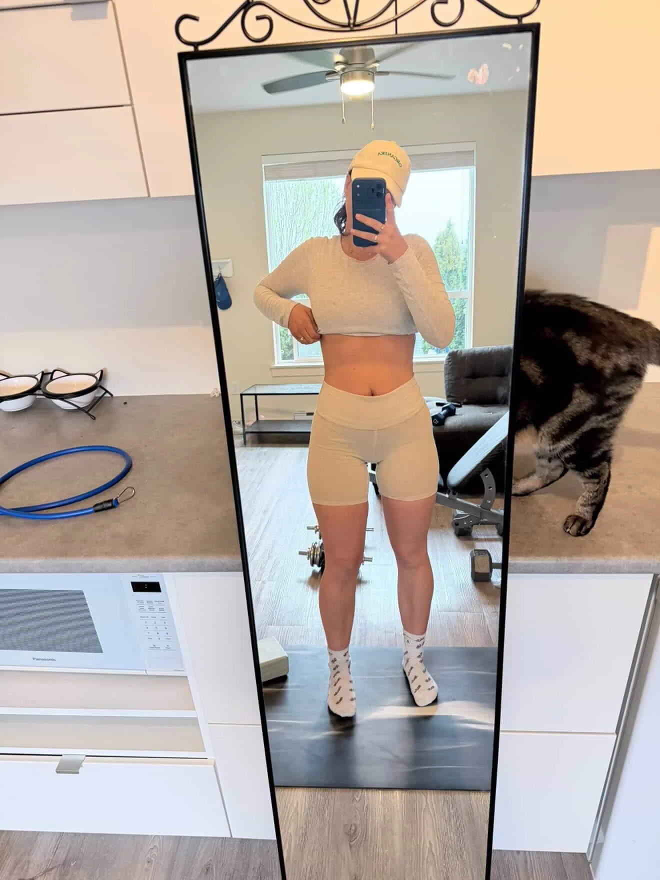 A woman taking a mirror selfie in a room with a microwave, dumbbells, and a cat in the background, wearing a long-sleeve crop top, shorts, and socks.