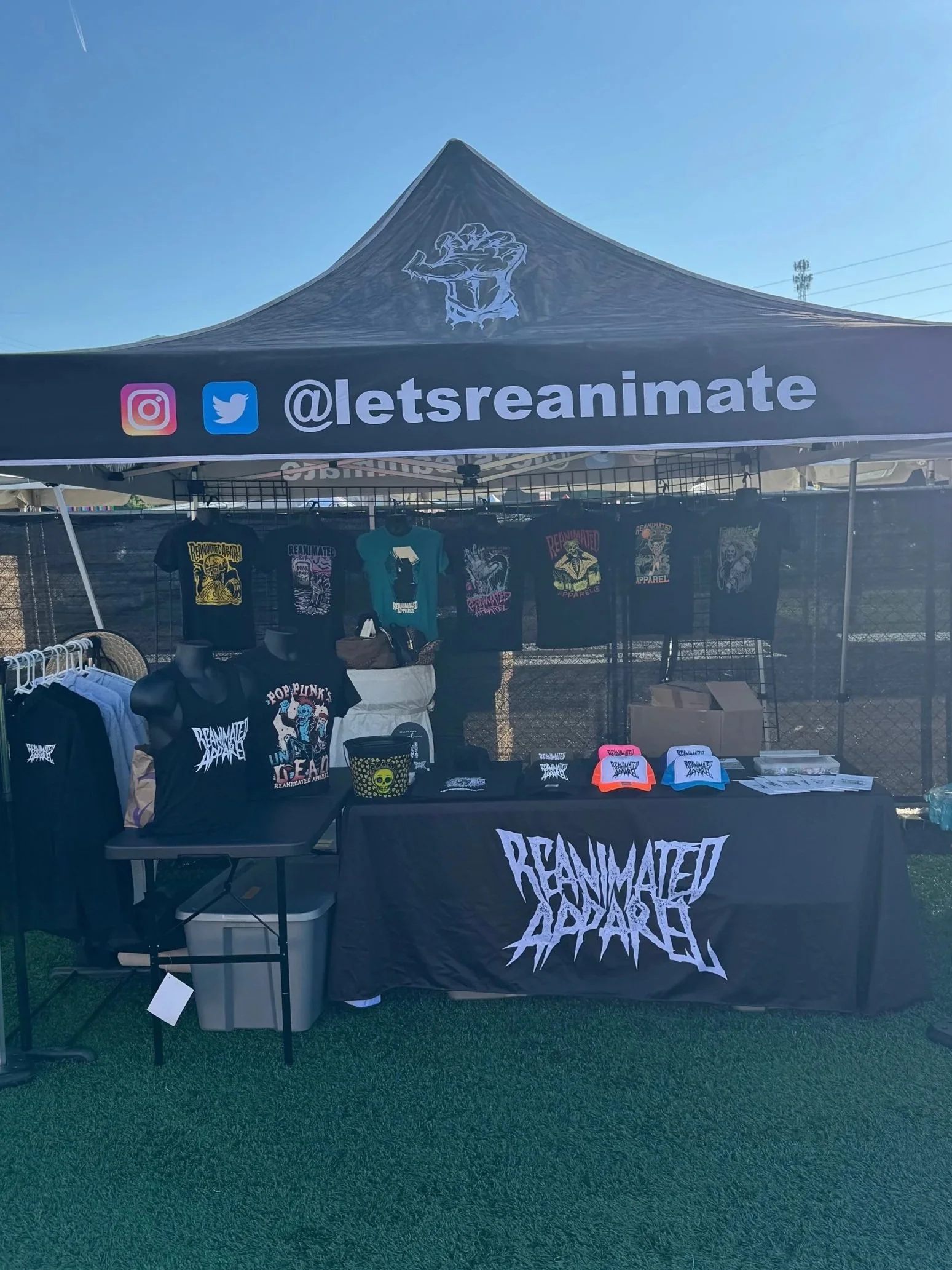A merchandise booth selling band or clothing brand items at an outdoor event, with a black canopy tent displaying the Instagram and Twitter handles '@letsreanimate'. The booth features t-shirts, hats, and other apparel with graphics and text, and a black tablecloth with 'Reanimated Apparel' printed on it.