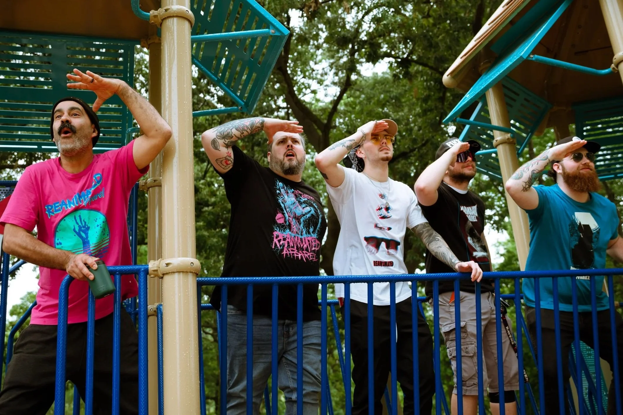 Group of five men standing on a playground, saluting or shading their eyes, with trees in the background.