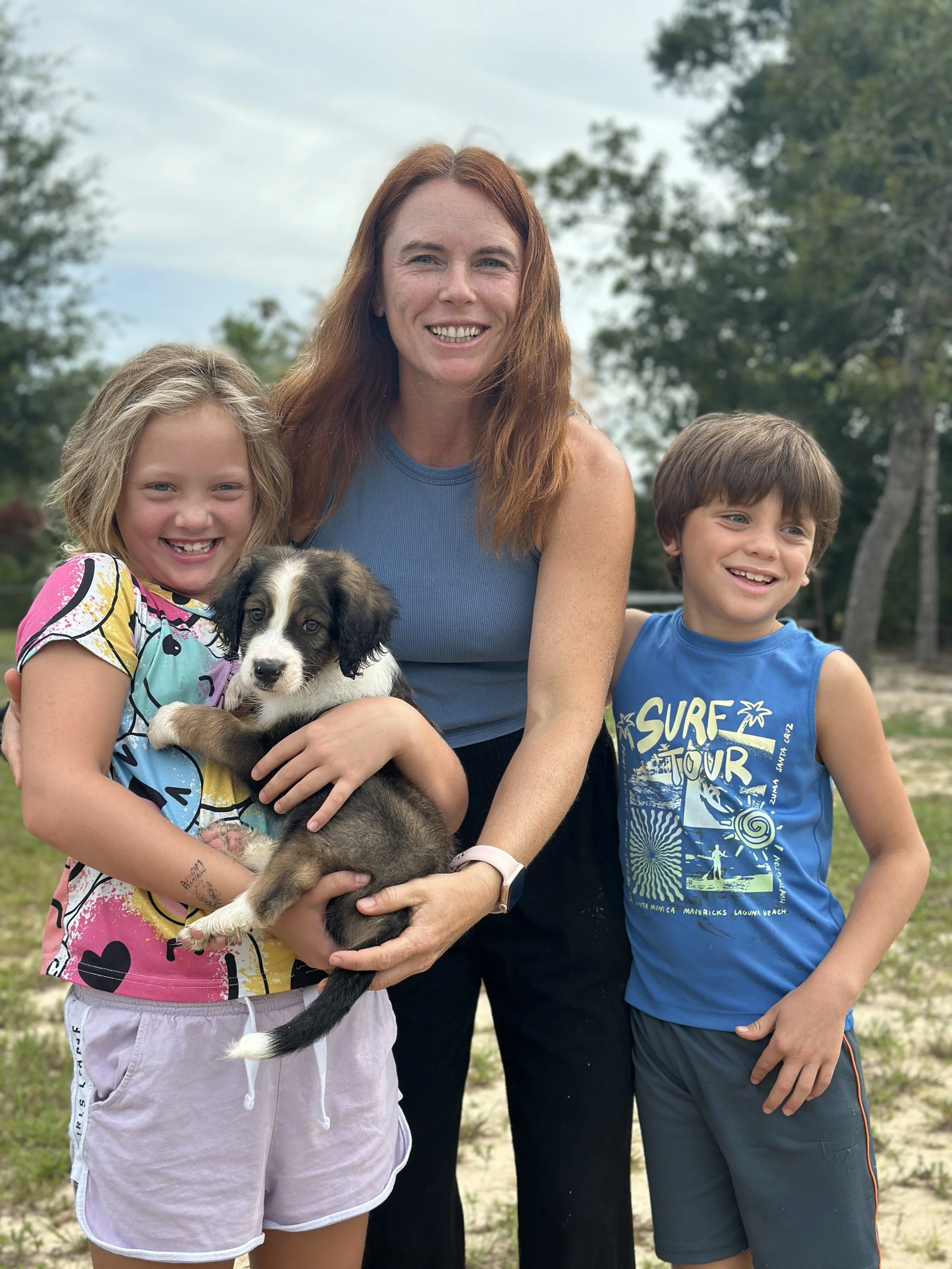 A woman and two children outdoors with trees in the background, smiling while holding a puppy.