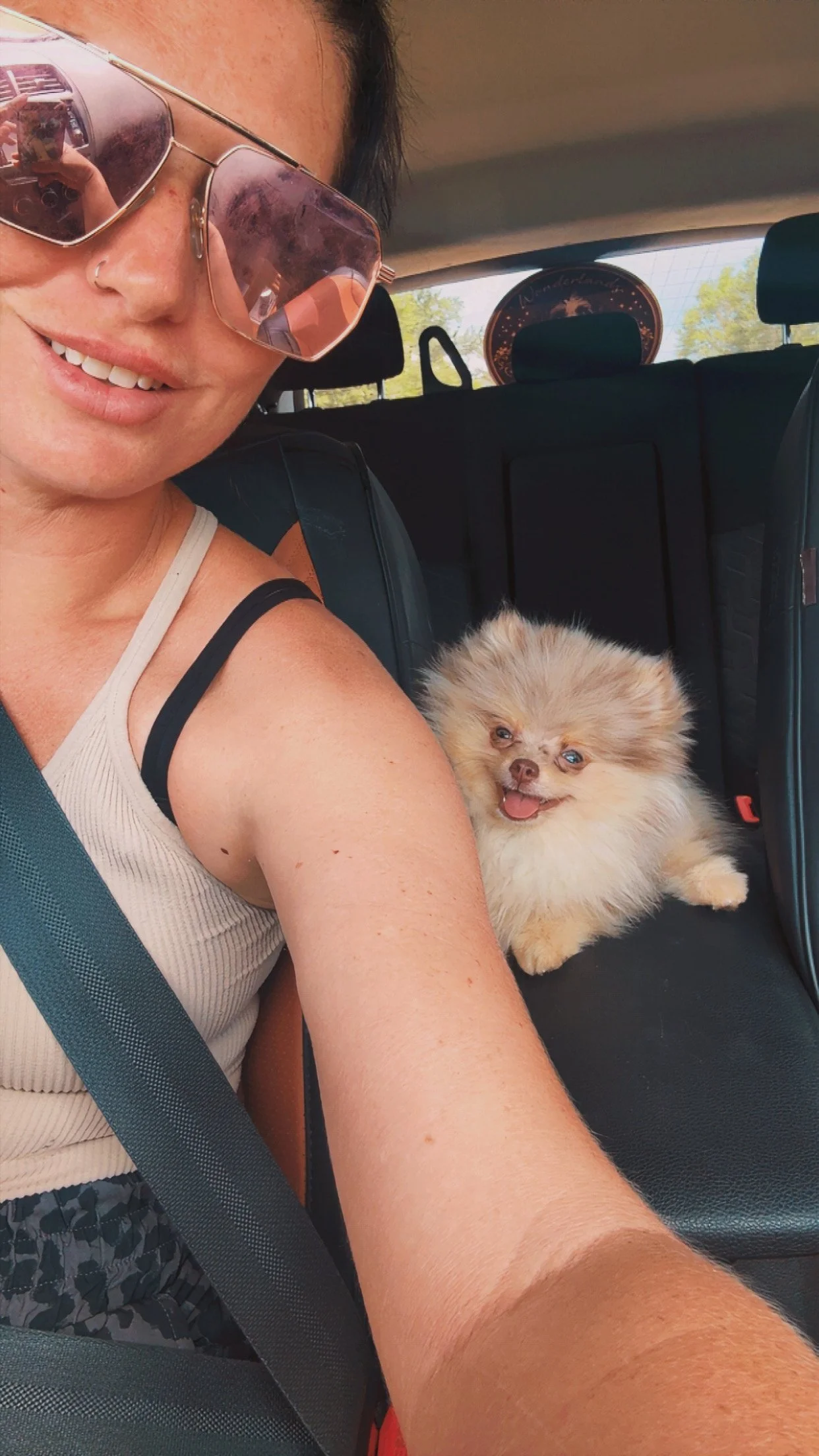 A woman wearing sunglasses and a striped tank top sitting in the car with a fluffy Pomeranian dog on her lap, both smiling.