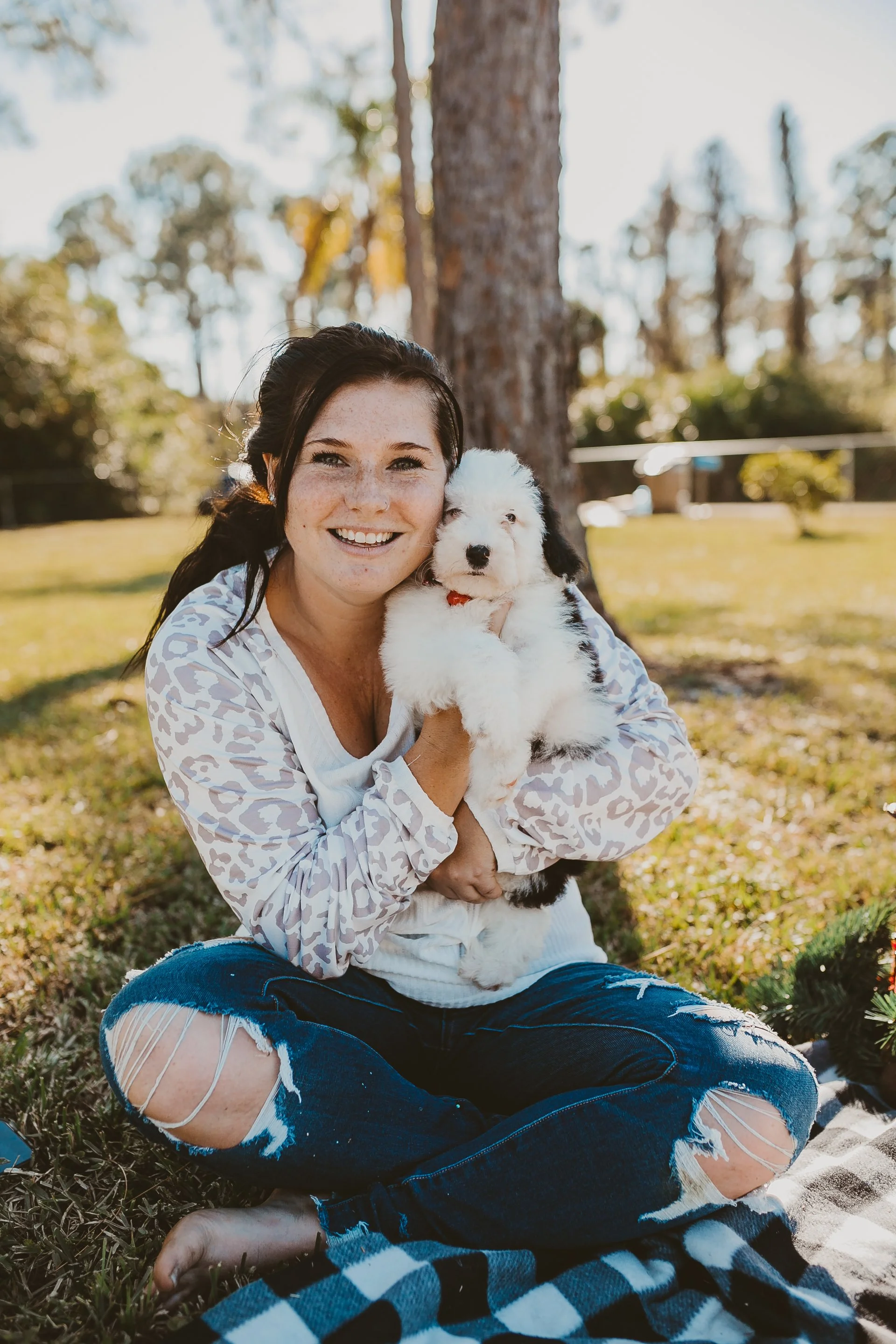 A woman sitting on the grass outdoors, holding a fluffy black and white puppy, with a large tree and a grassy field in the background.