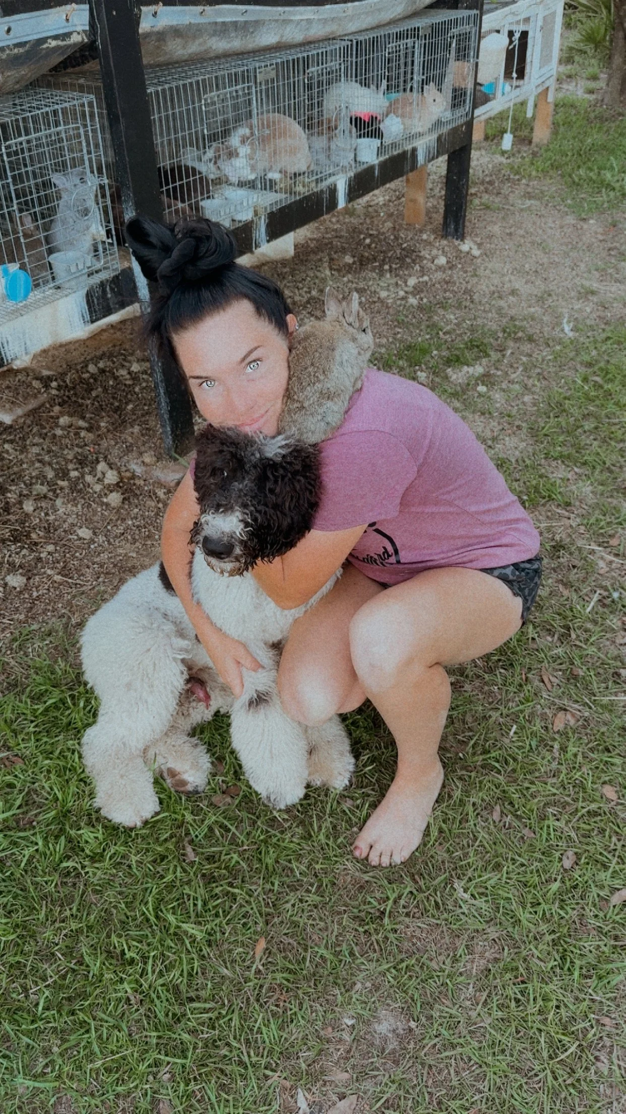 A woman hugging a large fluffy dog outdoors, with a cage of rabbits in the background.