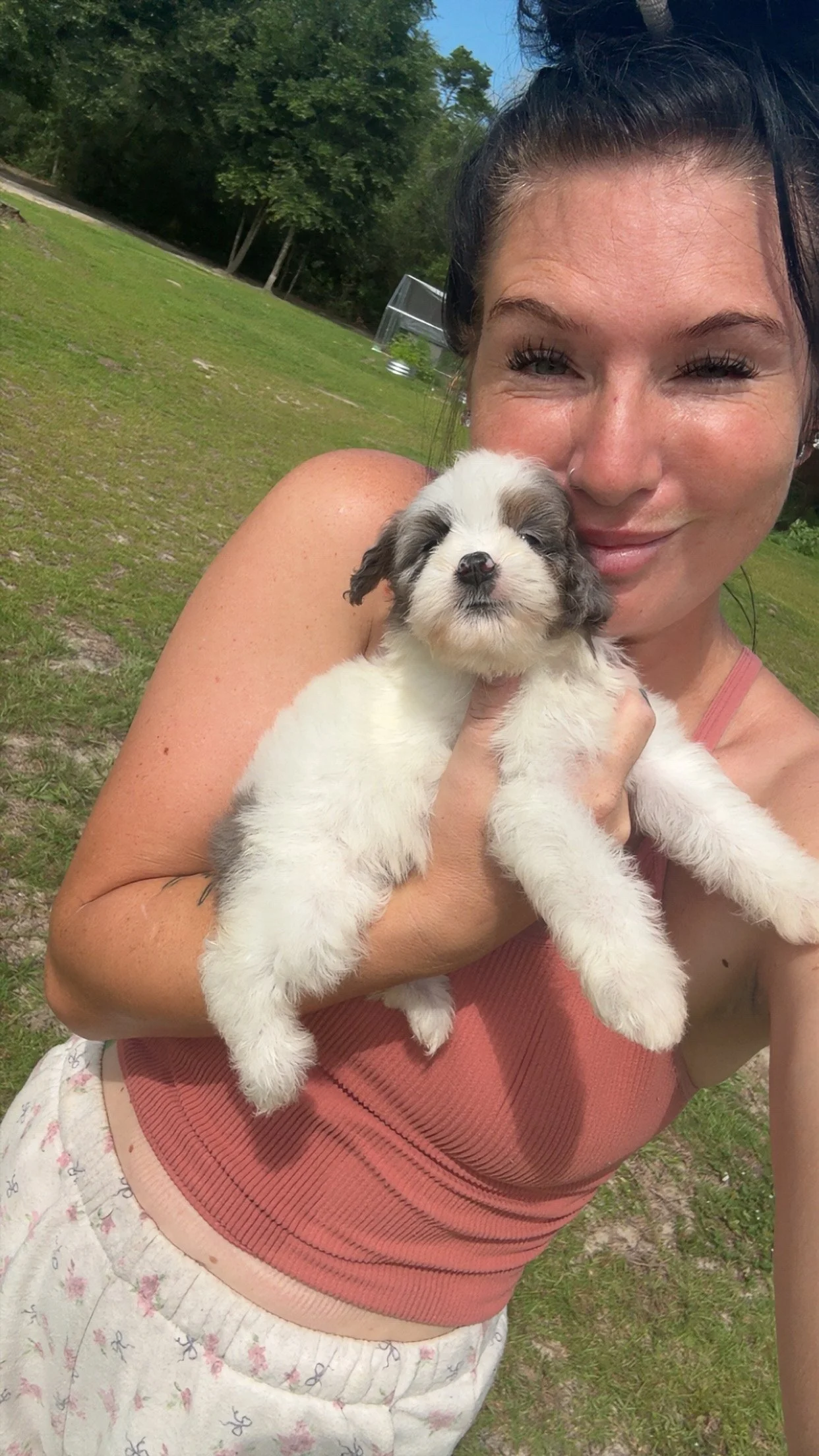 A woman with dark hair in a bun smiling and holding a cute white and gray puppy outdoors on grass with trees and a small greenhouse in the background.