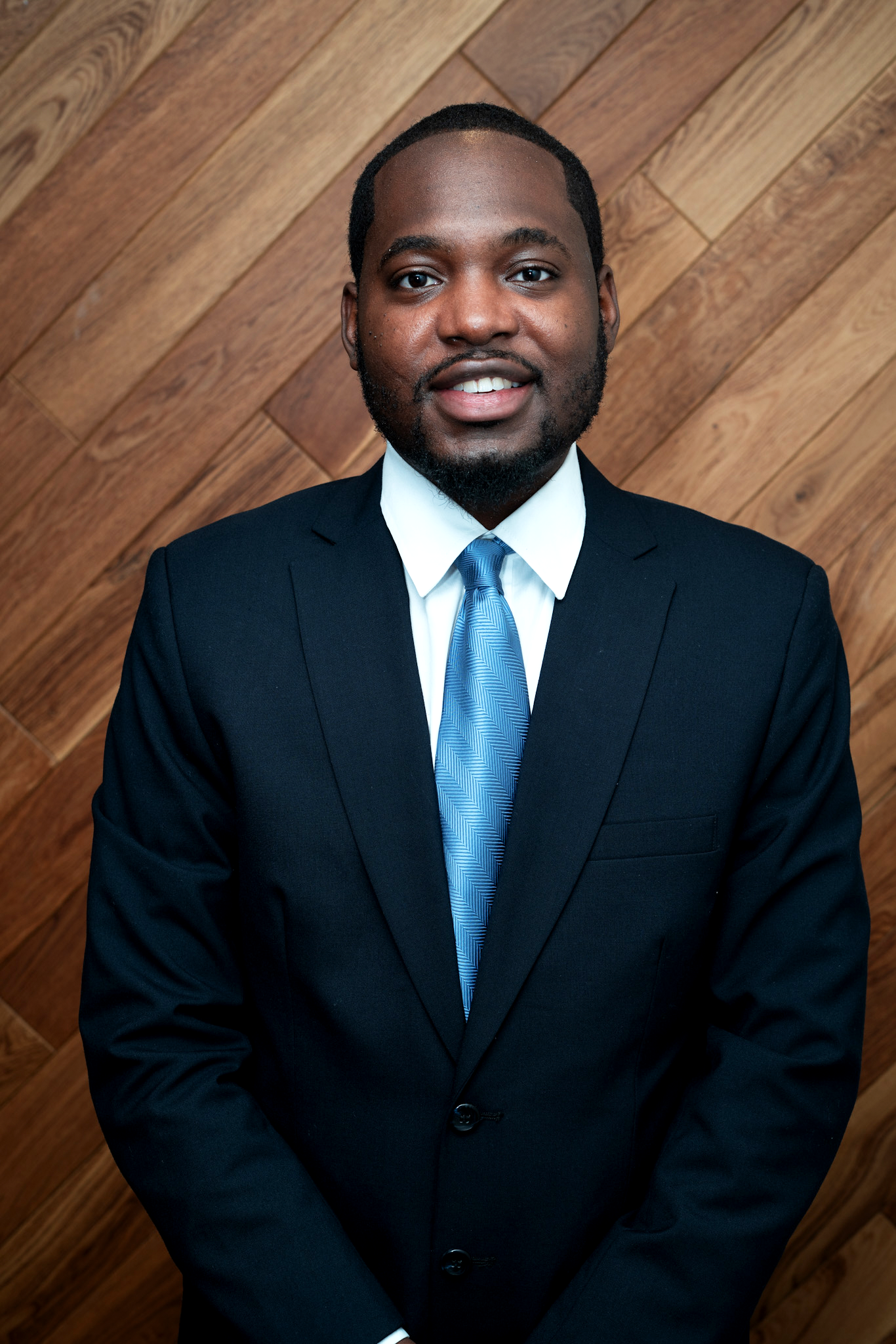 A man smiling in a black suit, white shirt, and blue striped tie, standing in front of a wooden wall.