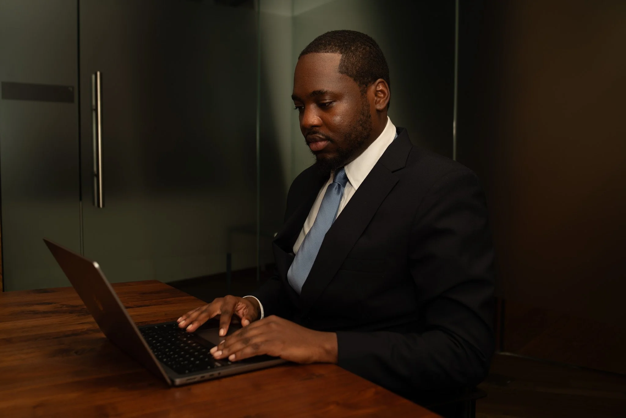 A man in a business suit working on a laptop at a wooden table in an office setting.