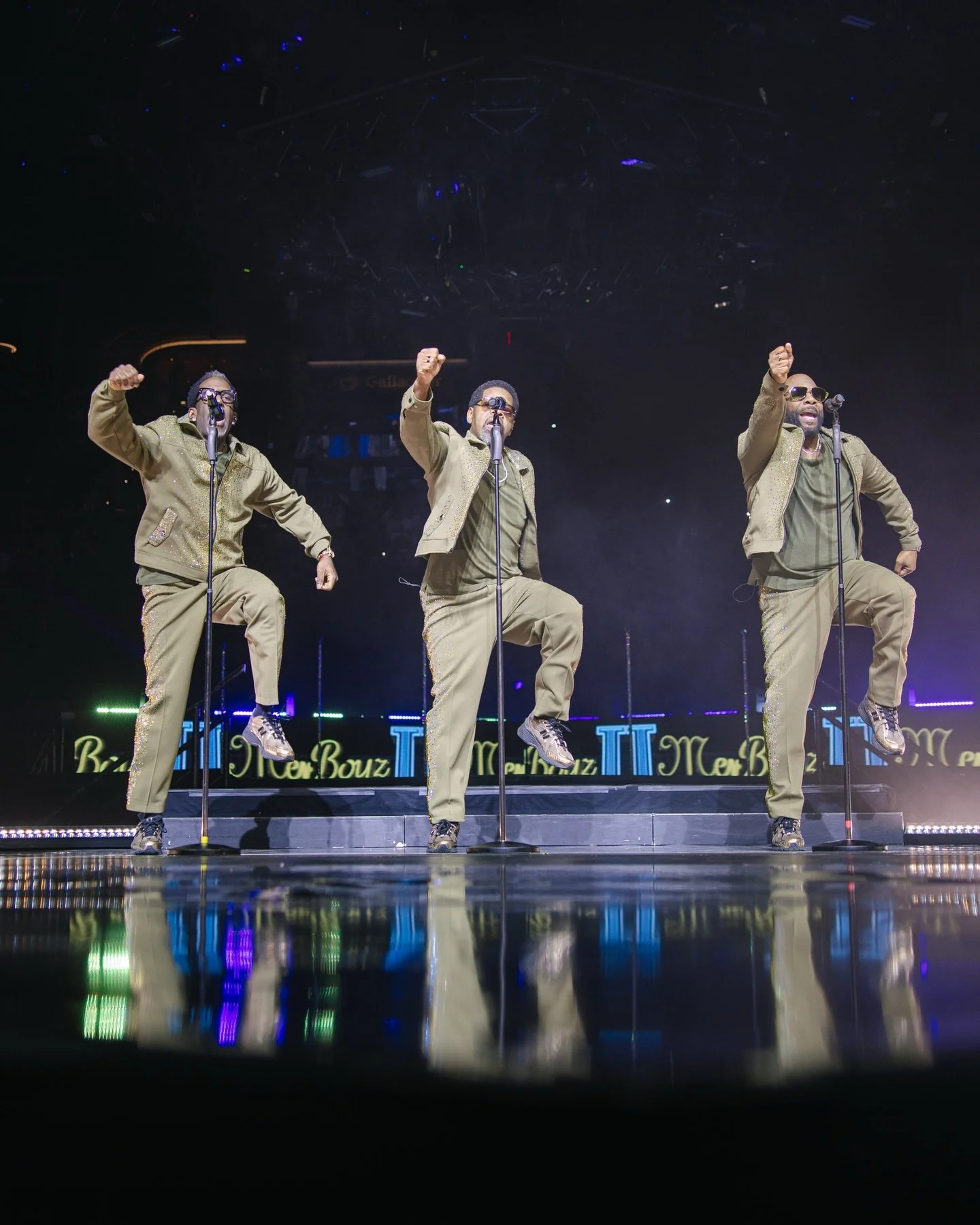 Three male performers wearing matching beige outfits on stage, singing into microphones with one leg raised in a synchronized dance move, illuminated by stage lights.