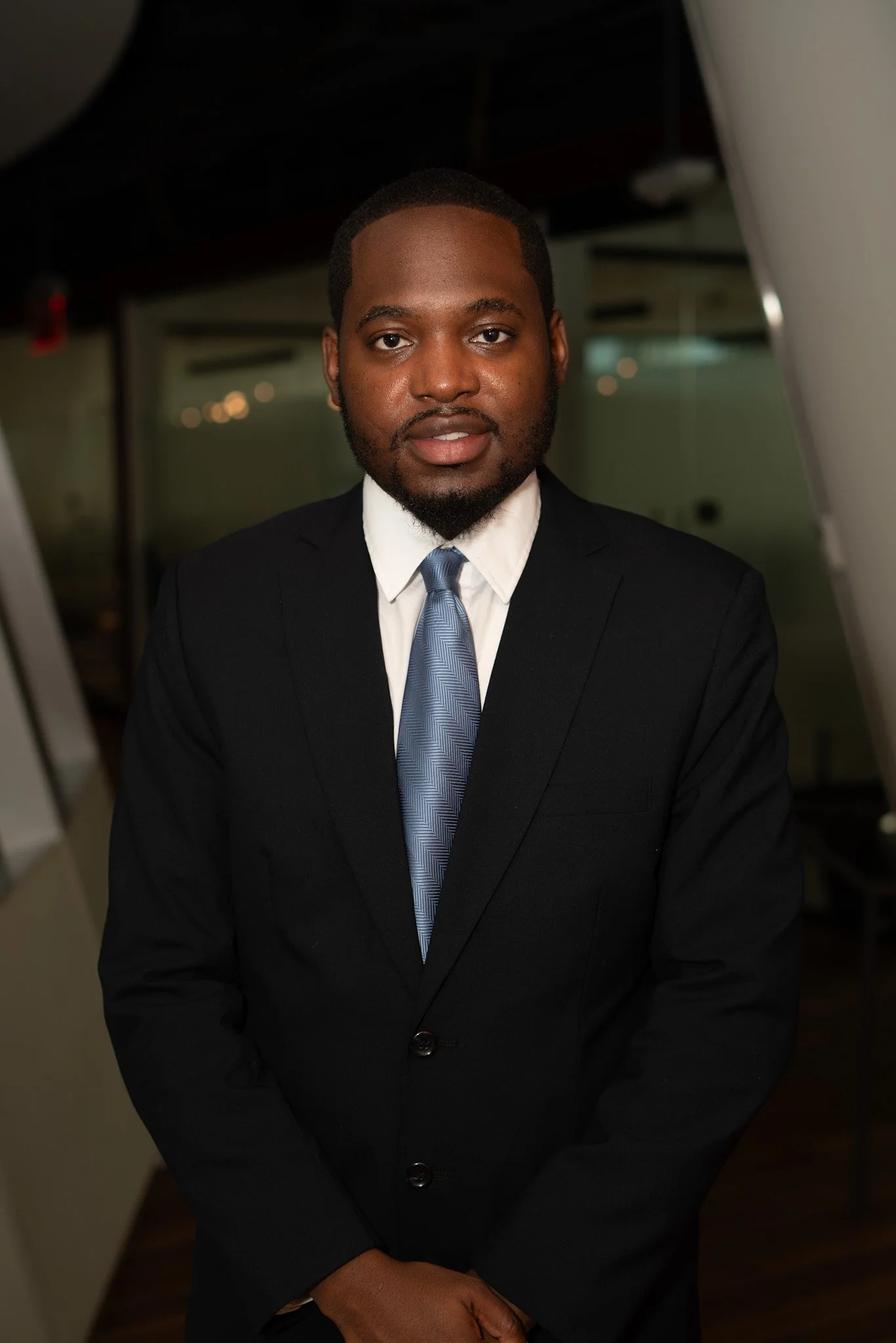 A man in a black suit with a white shirt and a blue tie standing indoors.