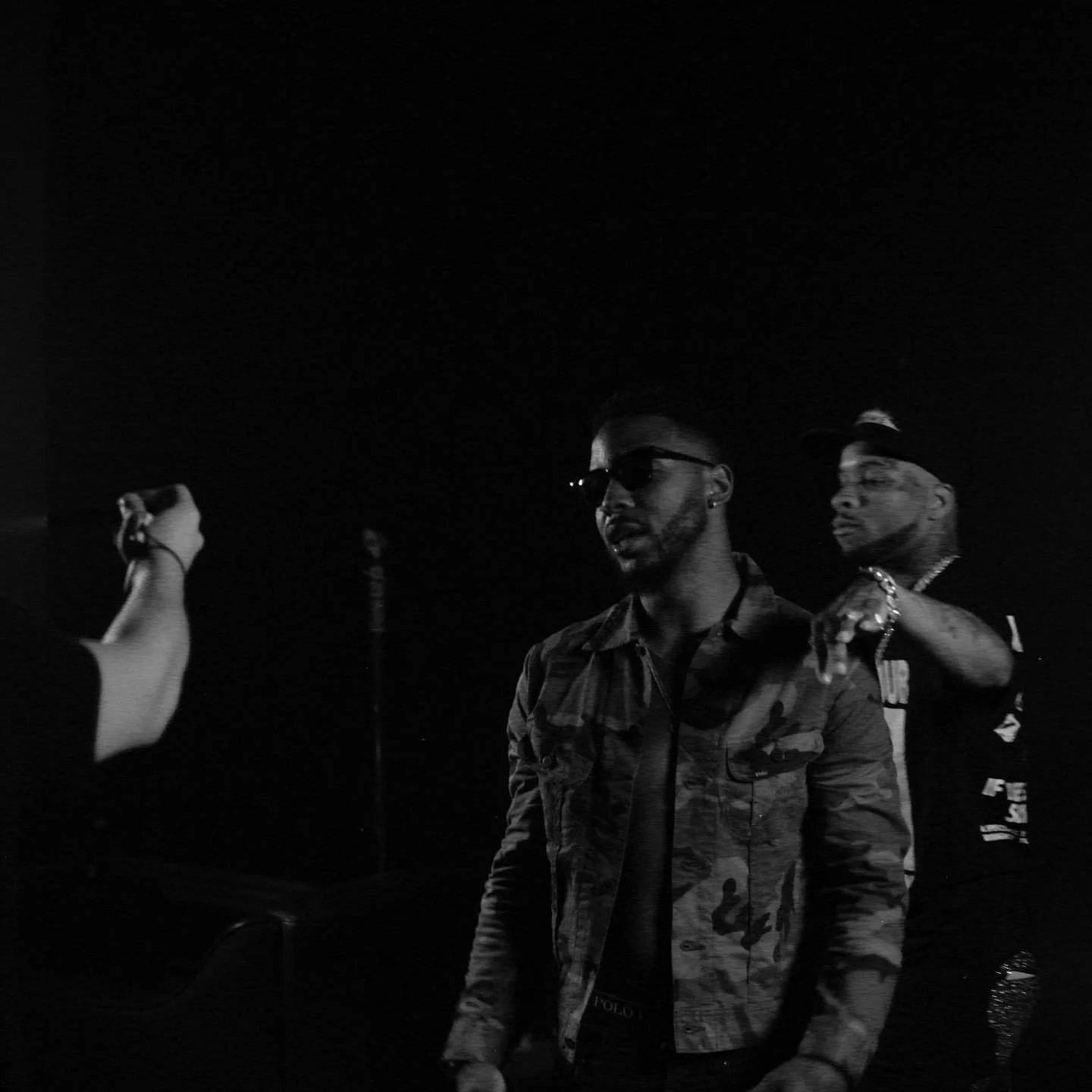 Two men in stylish outfits and sunglasses in a dark setting, with a person's hand holding a camera or phone in the foreground.