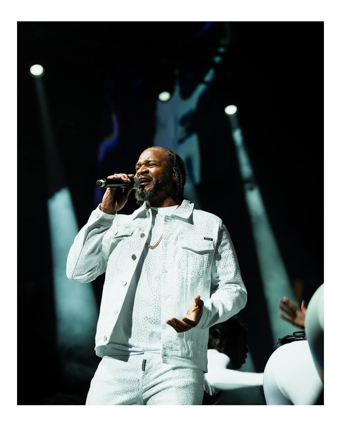 A male singer passionately performing on stage under bright spotlights, holding a microphone and dressed in a white jacket and pants.
