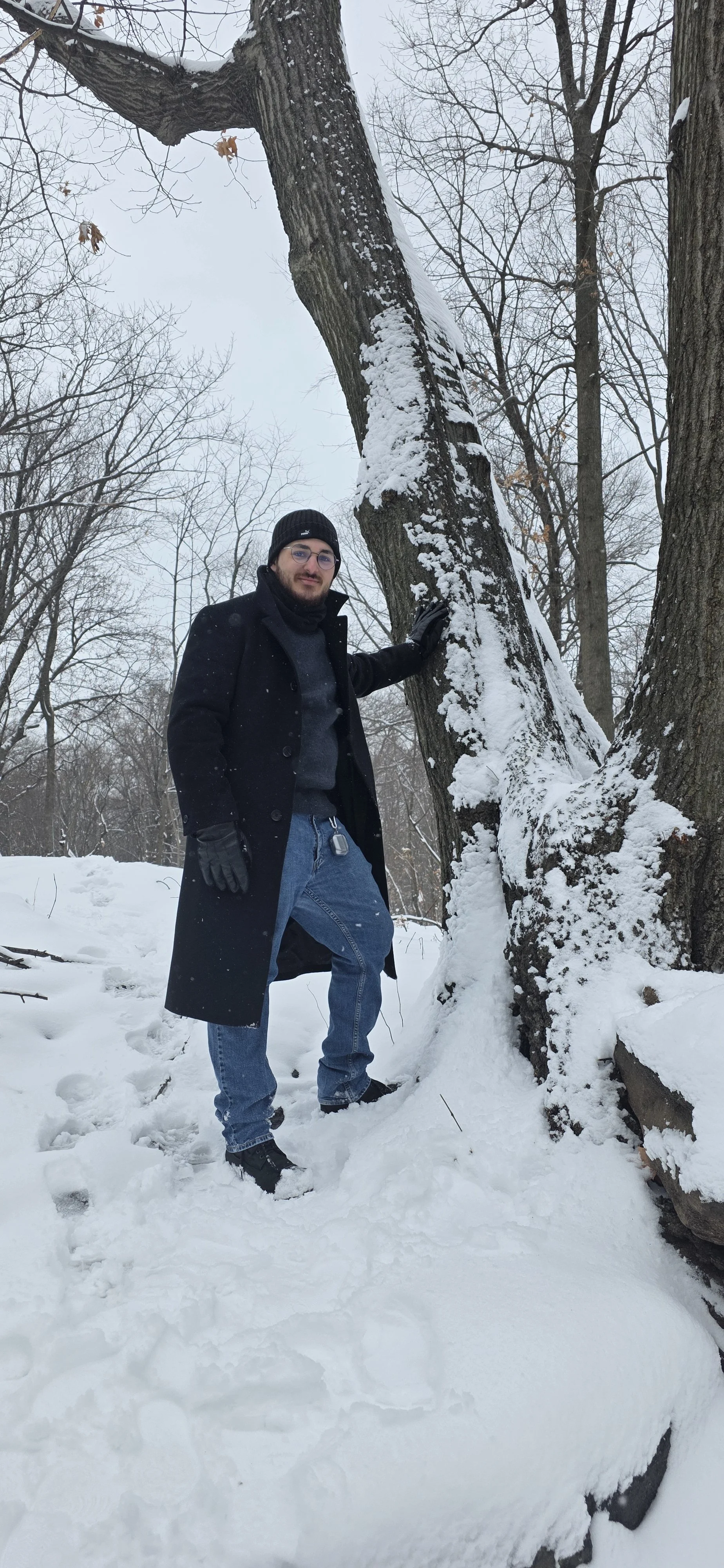A man dressed in winter clothing, including a black coat, black beanie, gloves, and glasses, standing in a snow-covered forest. He is touching a large, snow-covered tree trunk with his right hand.