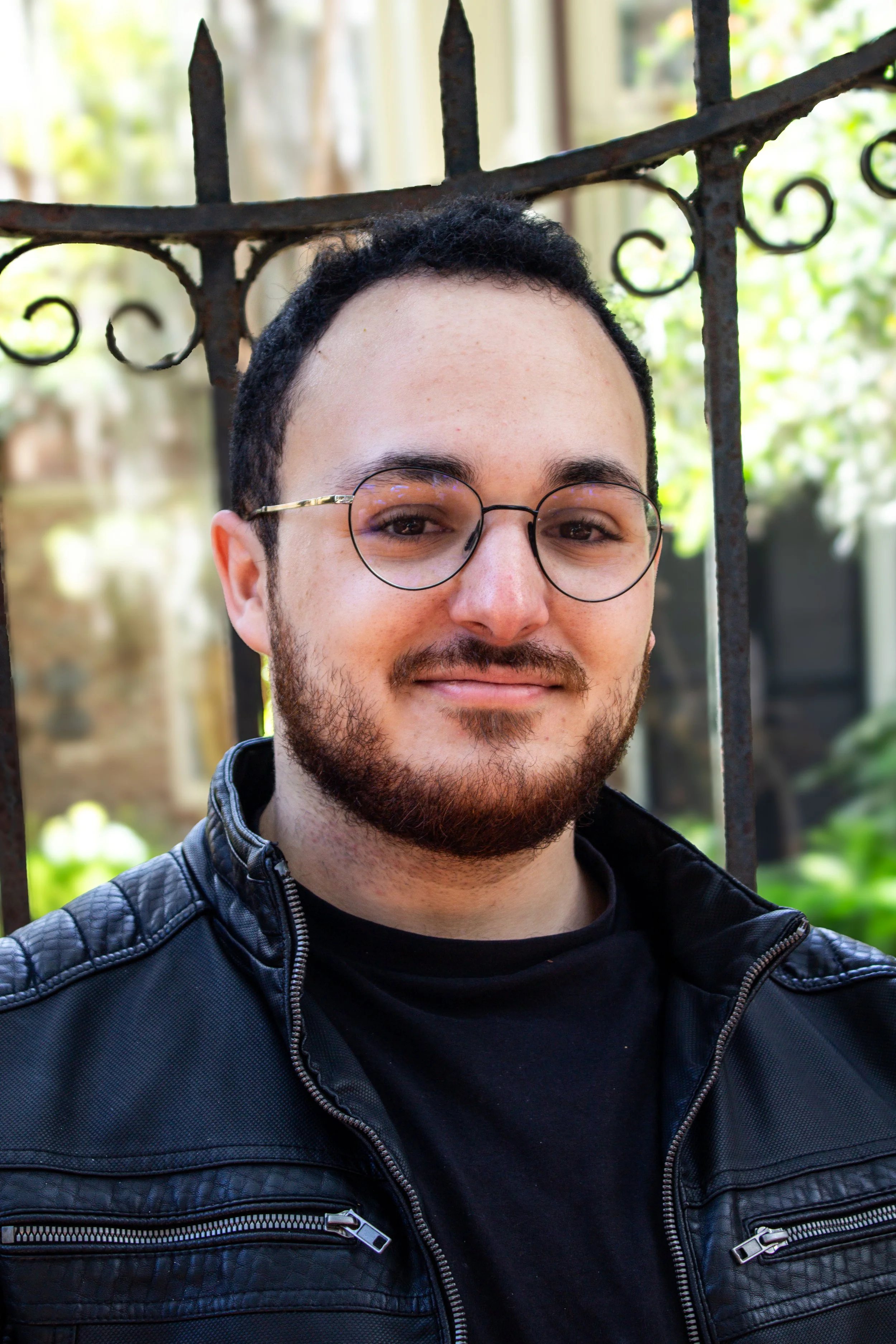 A young man with glasses and a beard smiling outdoors in front of a rusted iron fence with greenery in the background.