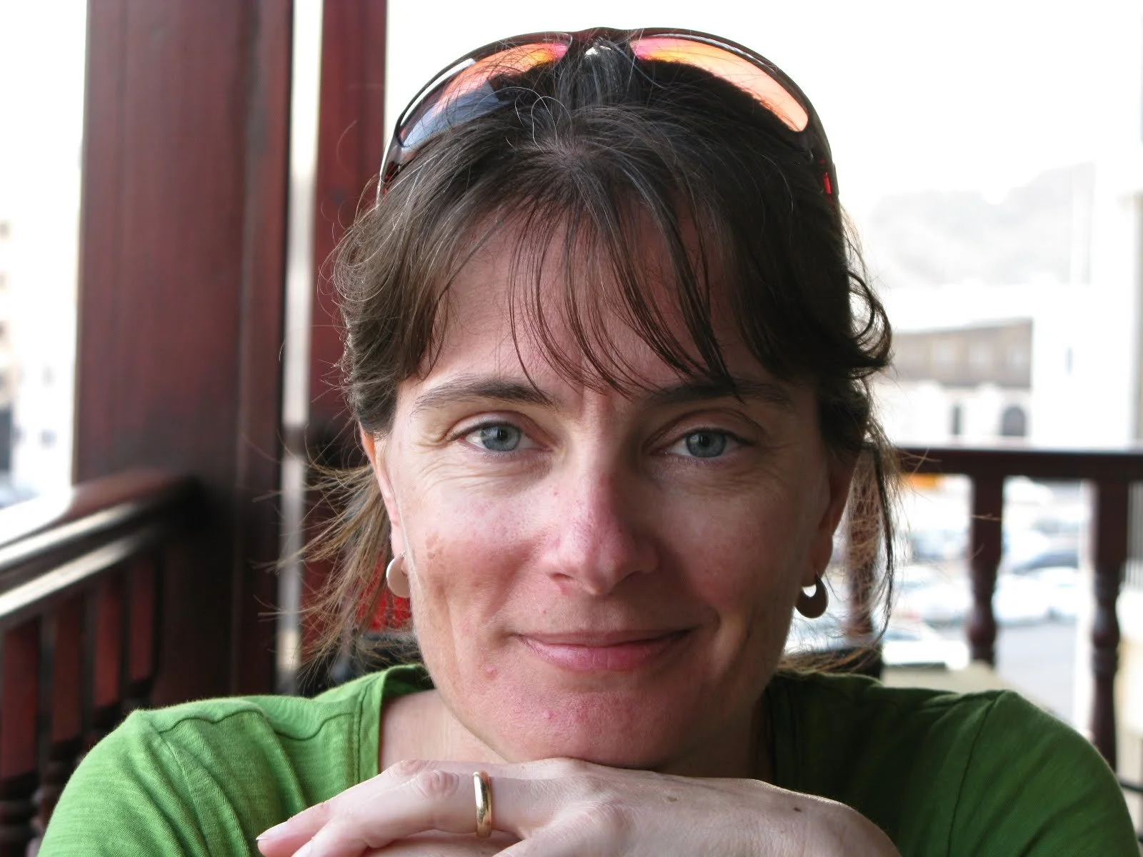 A woman with short brown hair, wearing glasses on top of her head and earrings, is smiling and leaning on her hands while sitting indoors near a window.