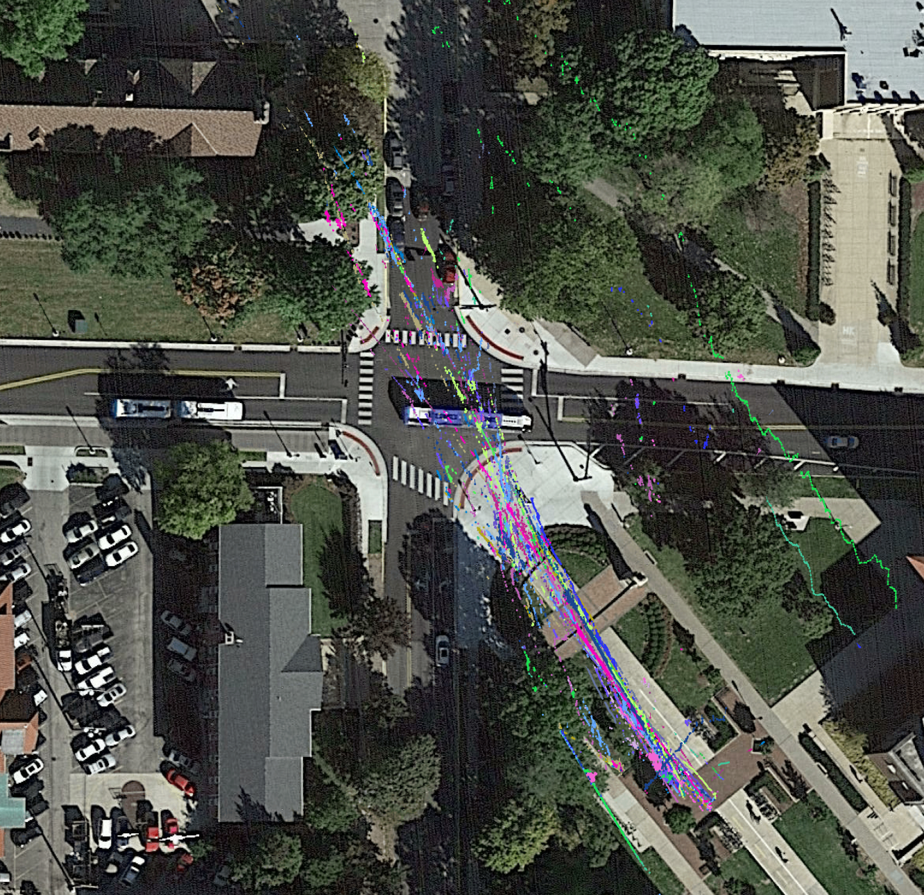 An aerial view of a city street intersection with various colored lines and markings, possibly indicating transit or traffic data, surrounded by trees, parked cars, and buildings.