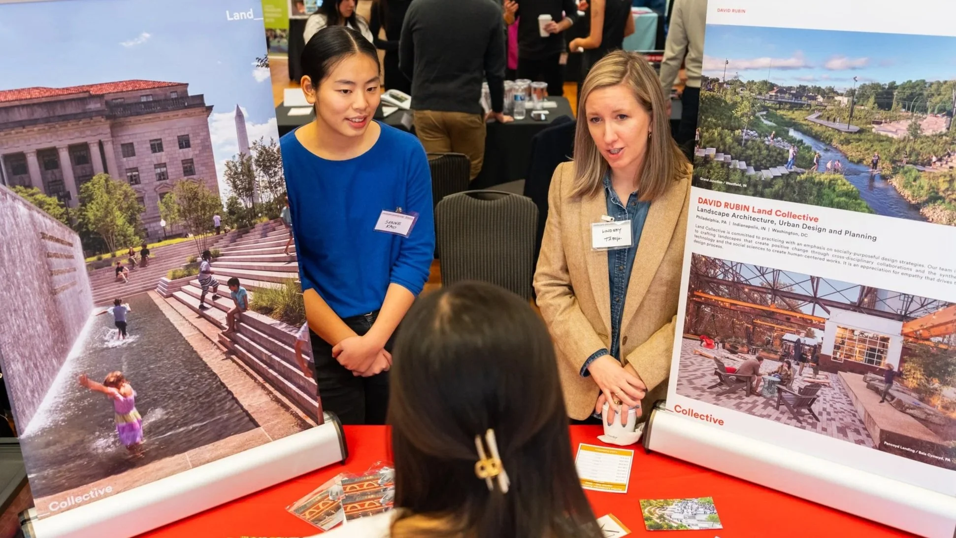 Two women are standing at a display booth, discussing landscape architecture projects. The booth features large posters of outdoor spaces with water features, steps, and green areas. One woman has blonde hair and is wearing a tan blazer, and the other has dark hair and is wearing a blue top. Several people are gathered around in the background at a conference or exhibition.