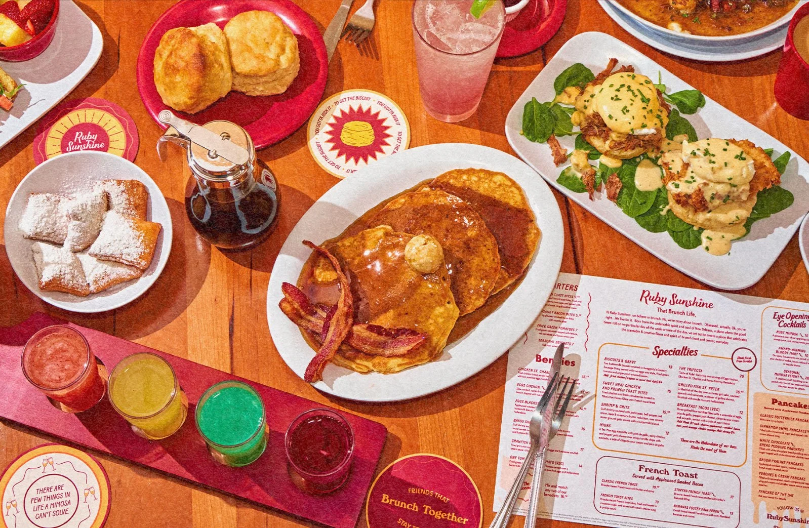 A breakfast table with various dishes: a plate of biscuits with powdered sugar, a glass of pink drink, a jar of syrup, a plate of biscuits and gravy, a large platter with eggs, bacon, and toast, a plate of breakfast sliders with eggs and bacon on brioche buns, and a class of colorful juices. A breakfast menu and small decorative coasters are also visible.