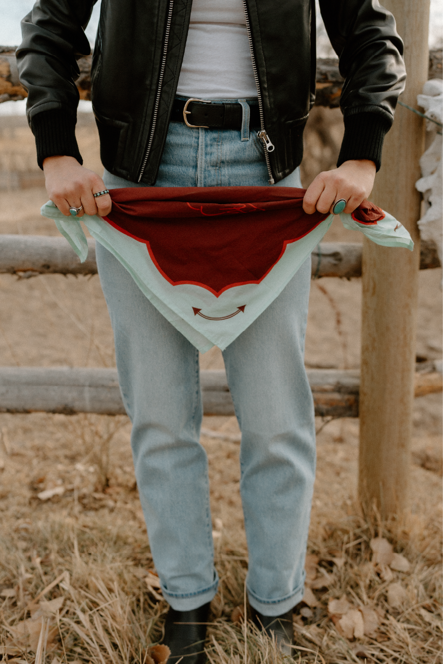 Person holding a bandana in front of their jeans, wearing a black leather jacket and rings, outdoors on a grass and dirt area.