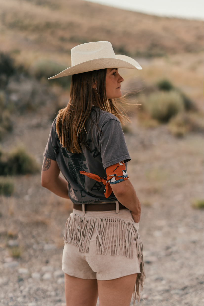 A woman standing outdoors in a dry, desert-like landscape, wearing a large white cowboy hat, a dark t-shirt, beige fringed shorts, and an orange bandana tied around her arm.