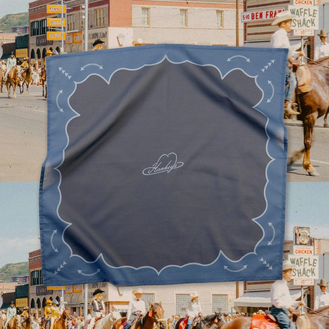 A blue bandana with white decorative border and the word "Hankey's" in cursive is in the foreground. In the background, there is a parade scene with people riding horses and storefronts, including signs reading "Waffle Shack" and "Ben Franklin".