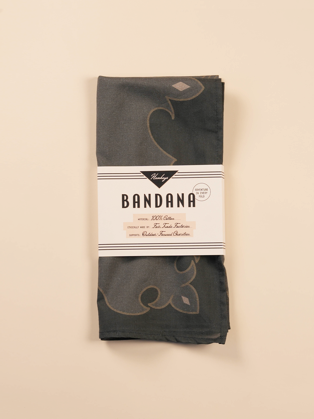 Folded dark gray bandana with a floral pattern, wrapped with a white band label that reads 'Hankeys BANDANA Material: 100% Cotton, Ethically made by: Fair Trade Factories, Supports: Outdoor-Focused Charities, Adventure in Every Fold'.