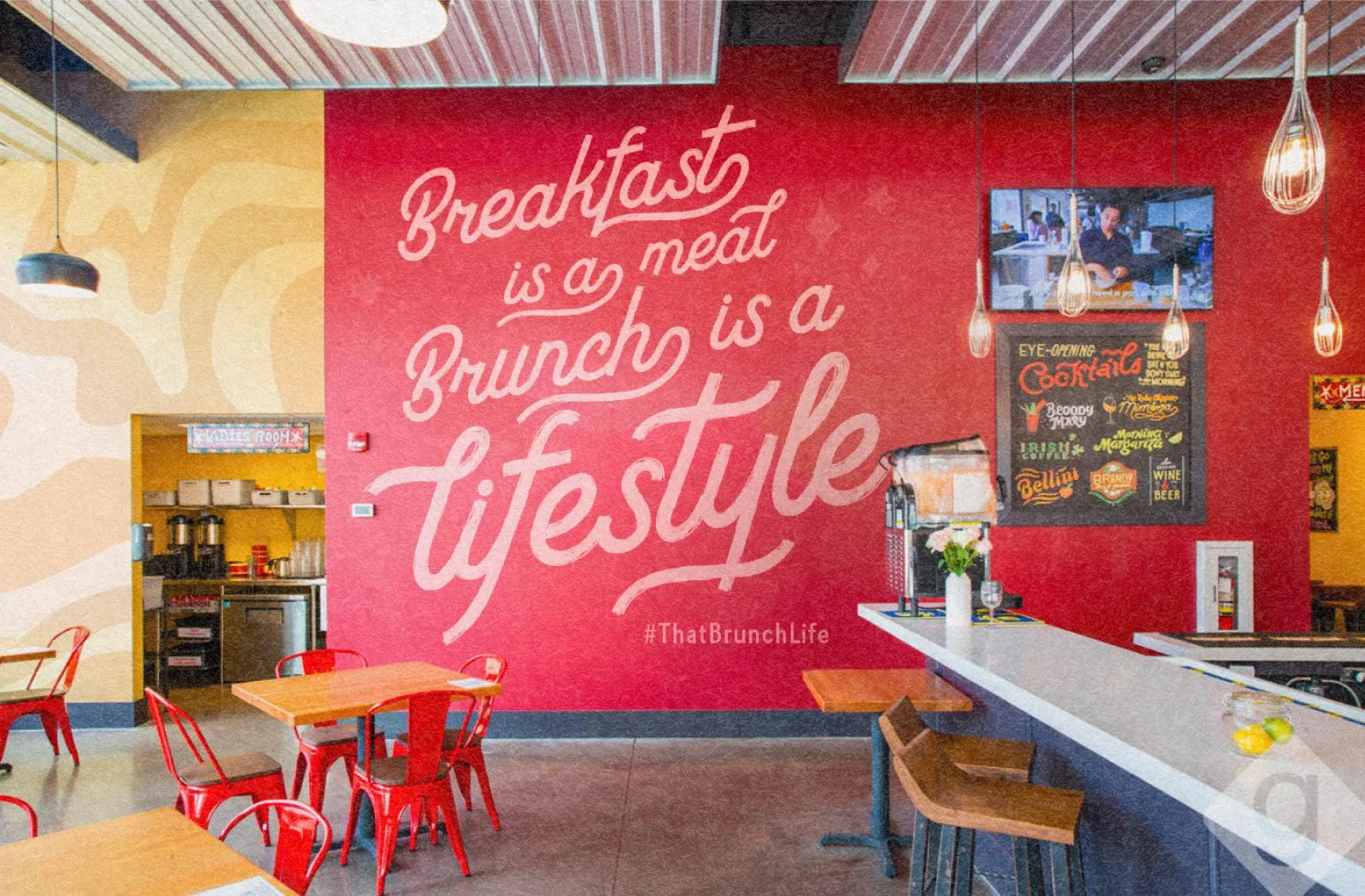 Interior of a restaurant with a red wall featuring a quote: 'Breakfast is a meat, brunch is a lifestyle' and a hashtag '#ThatBrunchLife', with tables, chairs, a coffee station, and a TV screen.