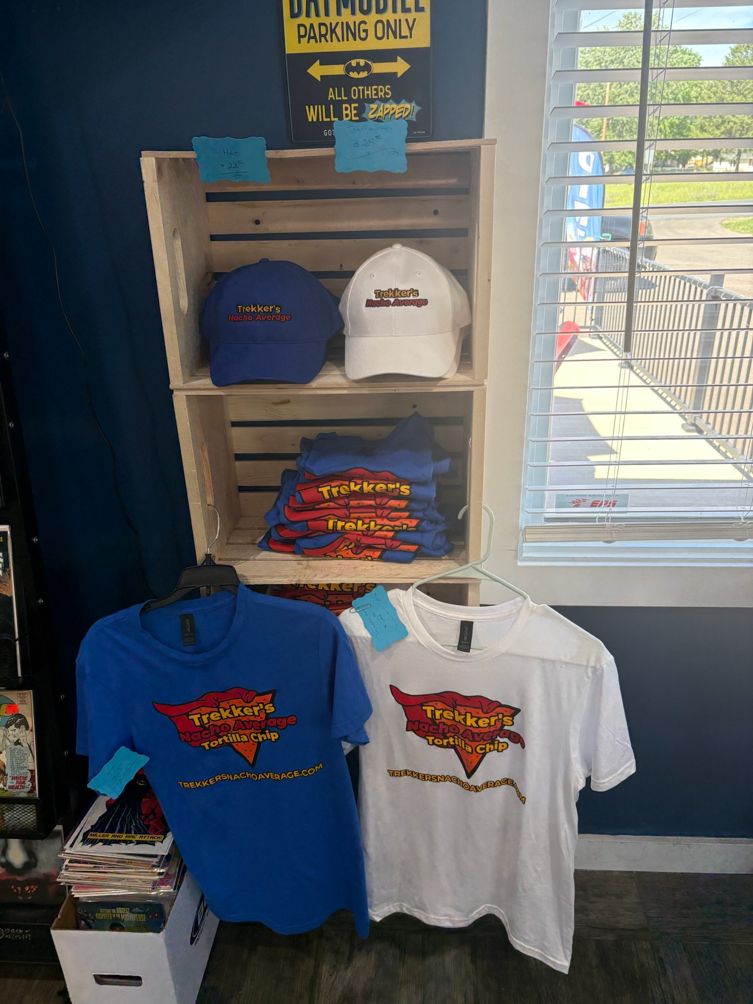 Display of T-shirts and caps with Nacho Average Tortilla Chips logo on shelves in store. The caps are blue and white, and the T-shirts are blue and white. Behind, there is a window with horizontal blinds and a parking lot outside.